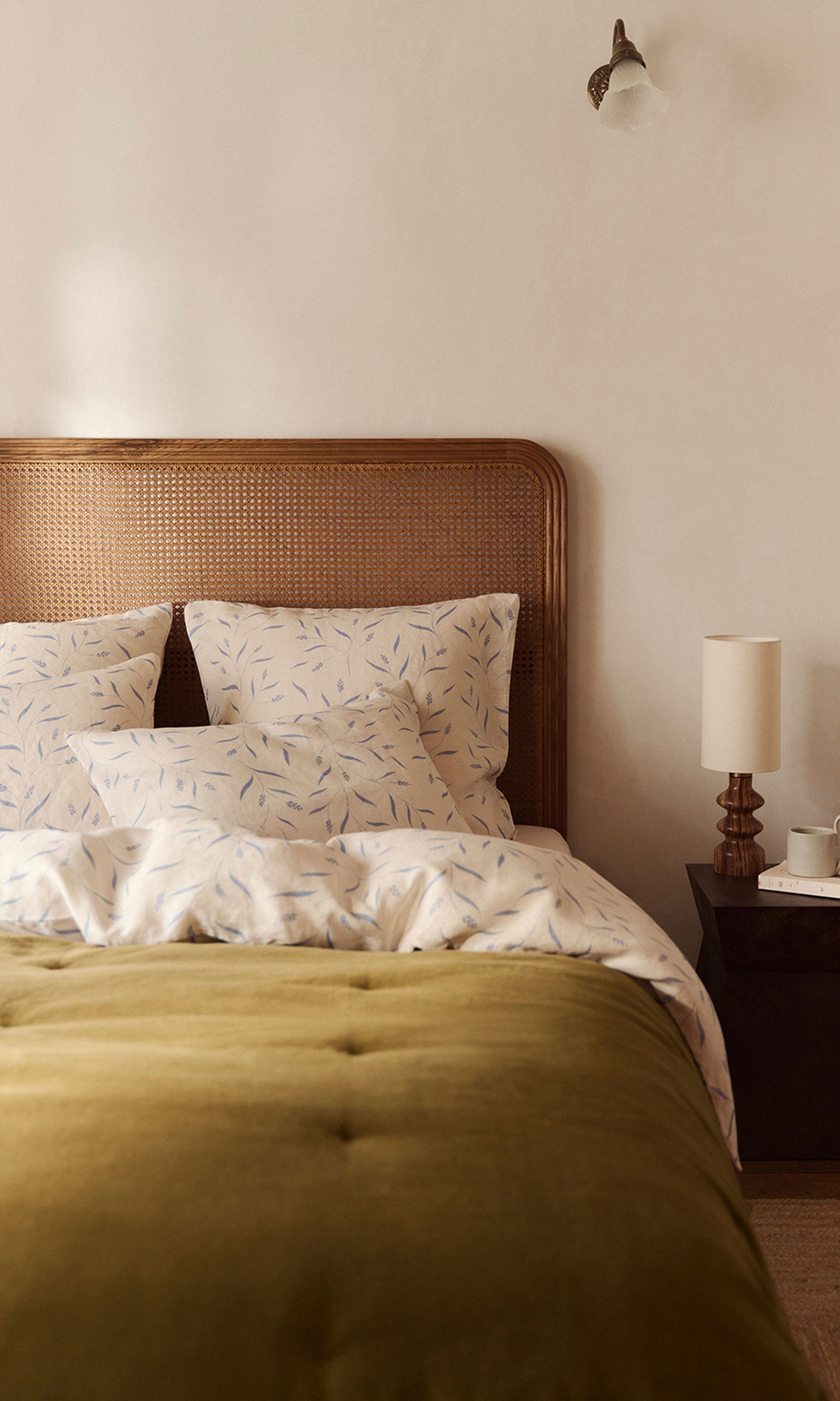 Une chambre douillette avec une tête de lit tressée, une parure de lit blanche et bleue à motifs de feuilles, une couverture vert olive, une table de chevet, une lampe et une applique murale.