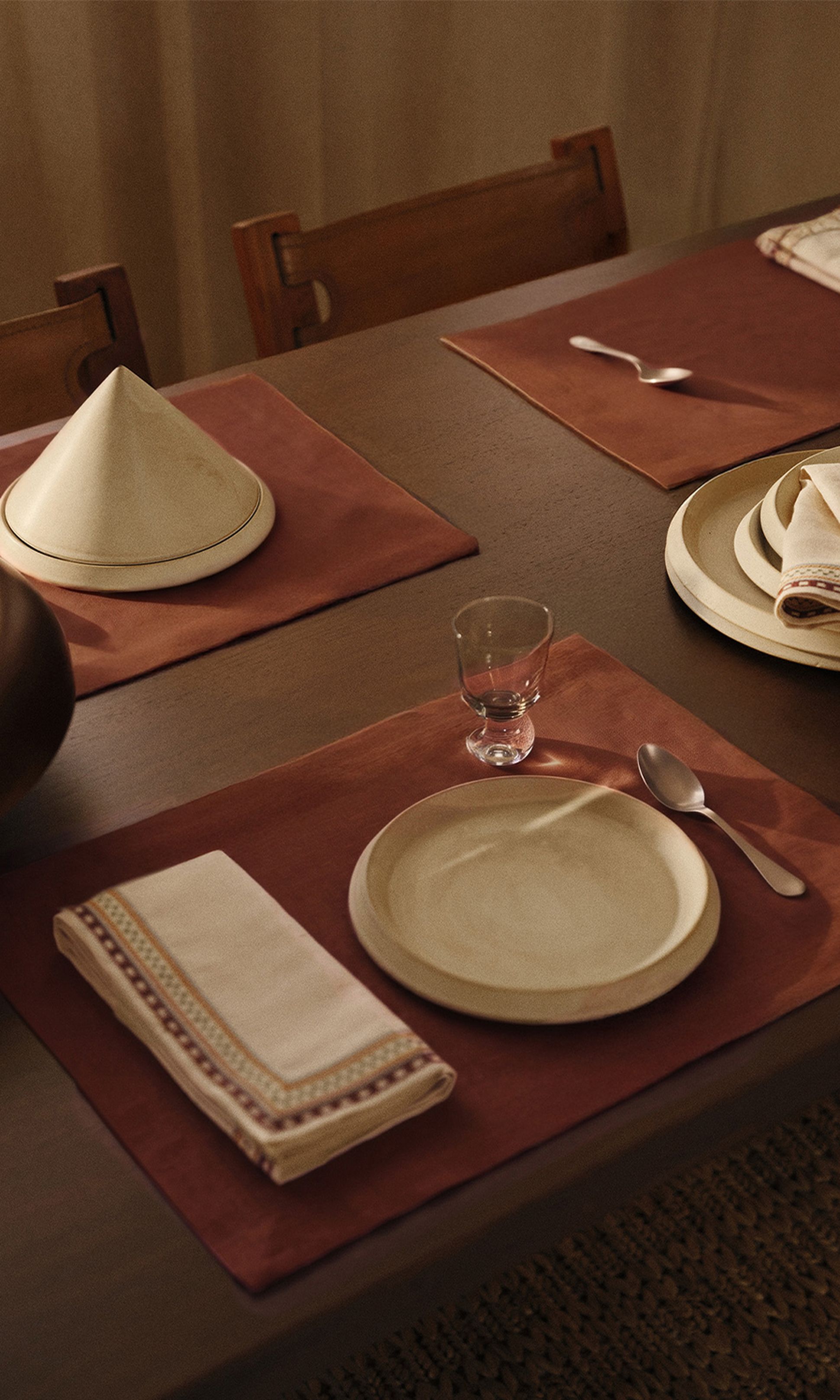 Une table élégamment dressée avec des assiettes beiges, une cuillère, un verre, une serviette pliée et un tajine posés sur des sets de table marron.