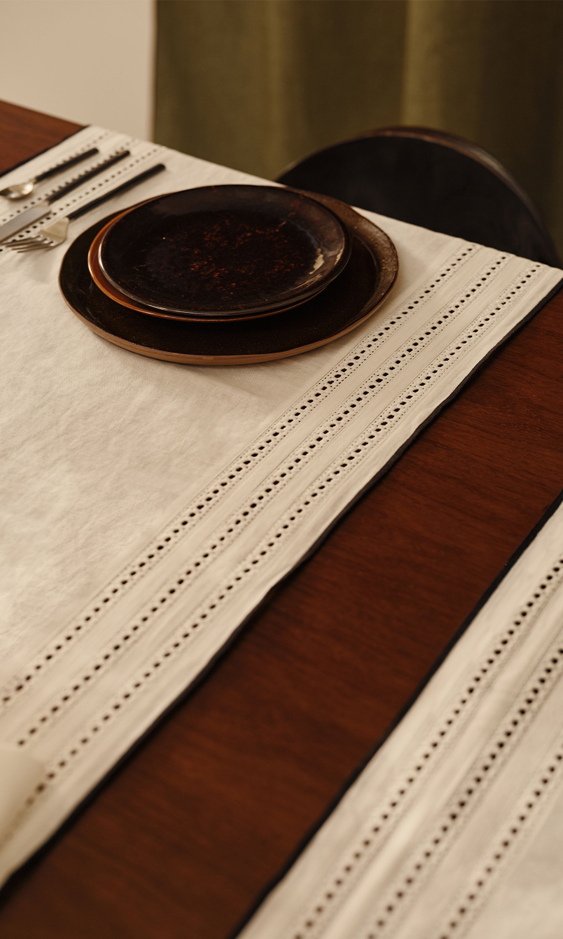 Une table élégamment dressée, avec deux assiettes foncées empilées sur un chemin de table blanc bordé de dentelle, les couverts à gauche et un plateau en bois en dessous.