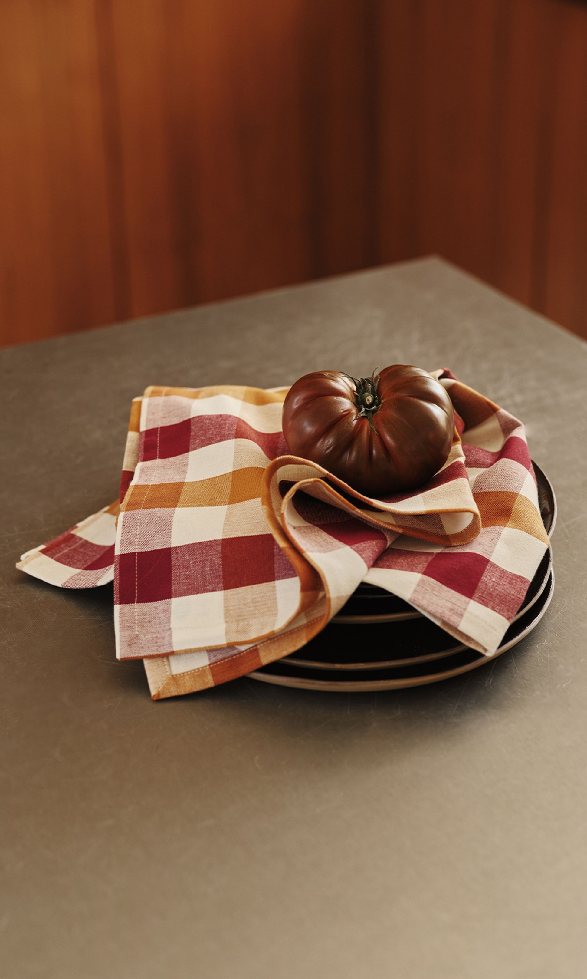 Une tomate foncée repose sur une pile d'assiettes recouverte d'une serviette à carreaux rouges, orange et blancs, posée sur un plan de travail.