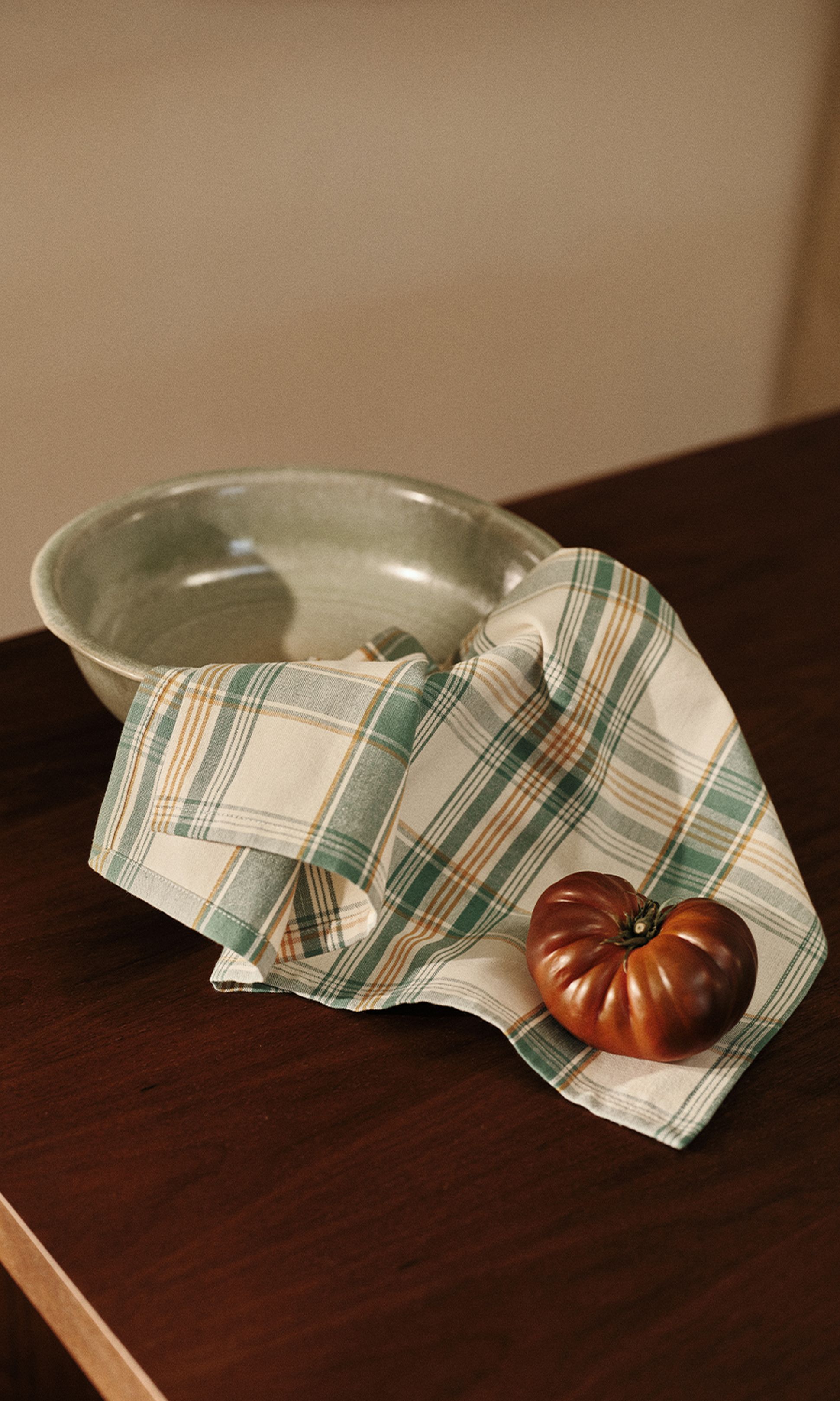 Un bol en céramique verte et un torchon rayé sur lequel repose une tomate, posés sur une table en bois.