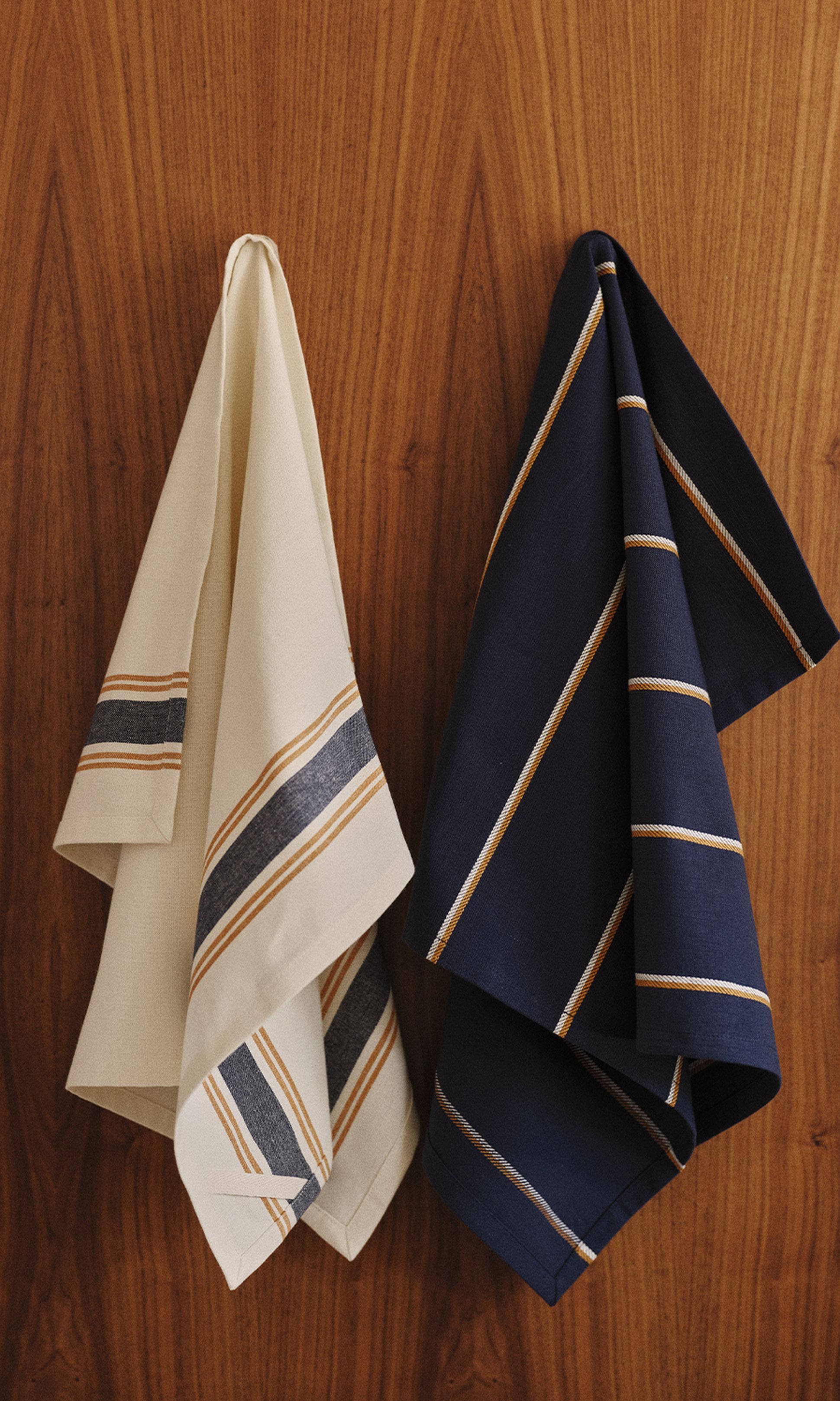 Deux torchons rayés, l'un beige et l'autre bleu marine, suspendus à un mur en bois.