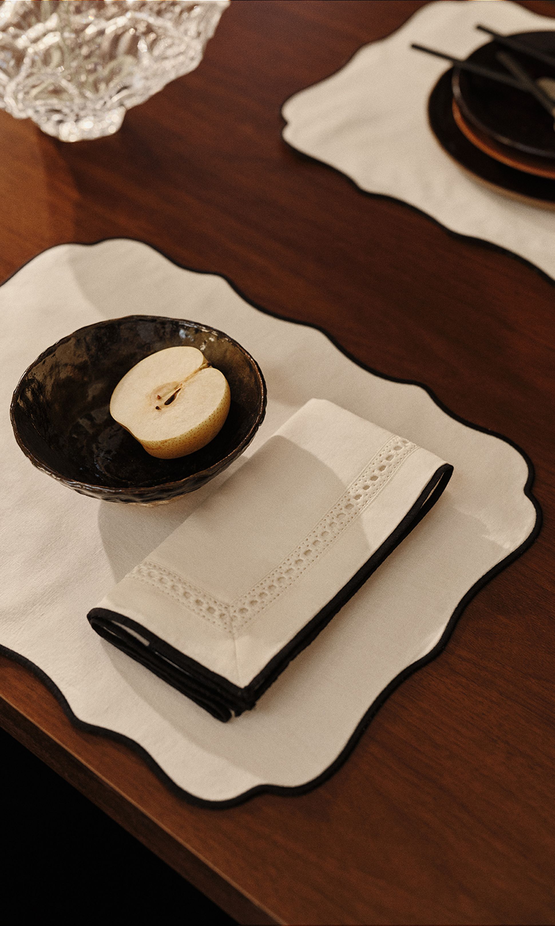 Une pomme coupée en deux repose dans un bol foncé posé sur un set de table festonné, avec une serviette pliée à côté, sur une table en bois.