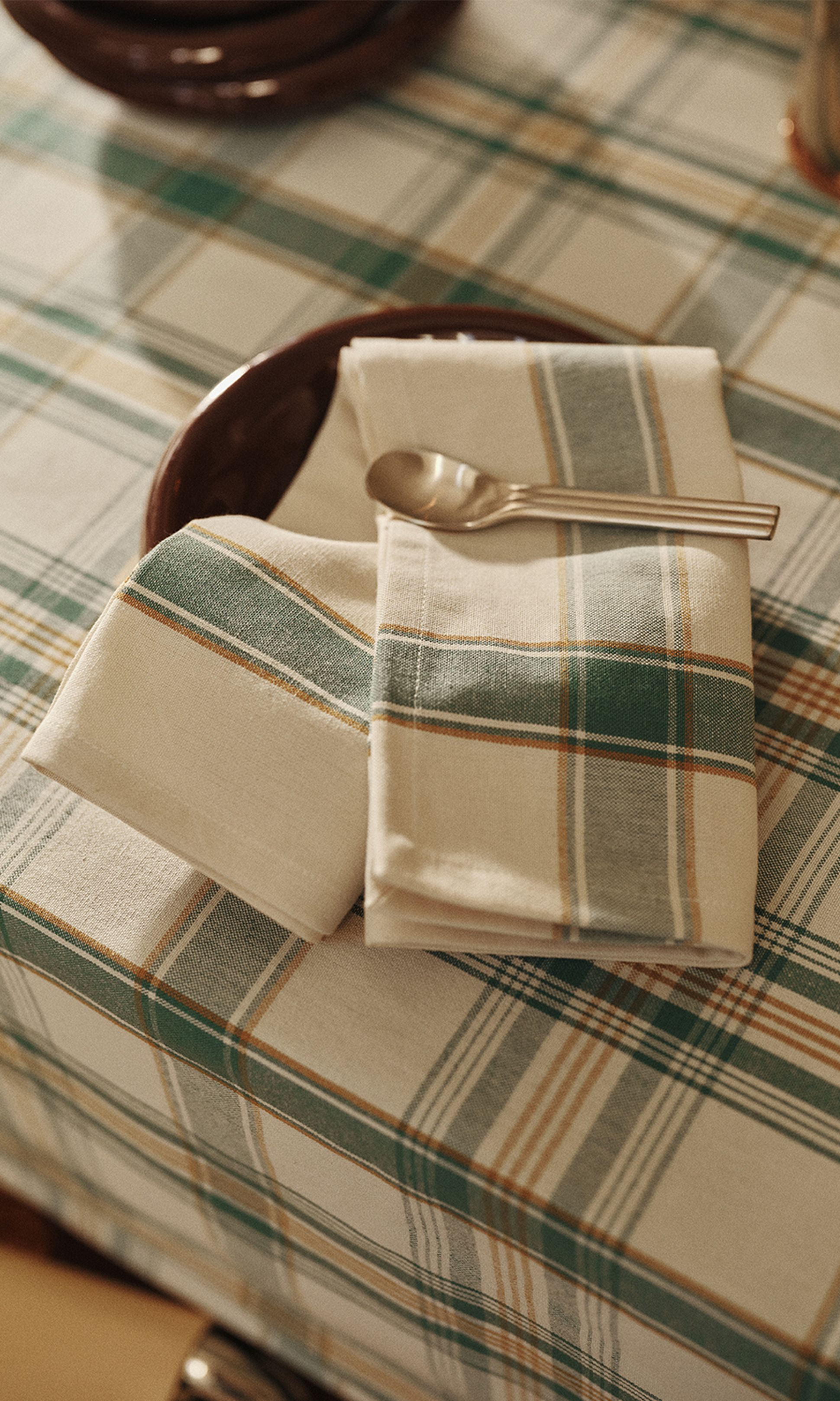 Une nappe à carreaux, accompagnée d'une serviette et d'une cuillère assorties posées sur une assiette marron, créant une table à l'ambiance chaleureuse et rustique.