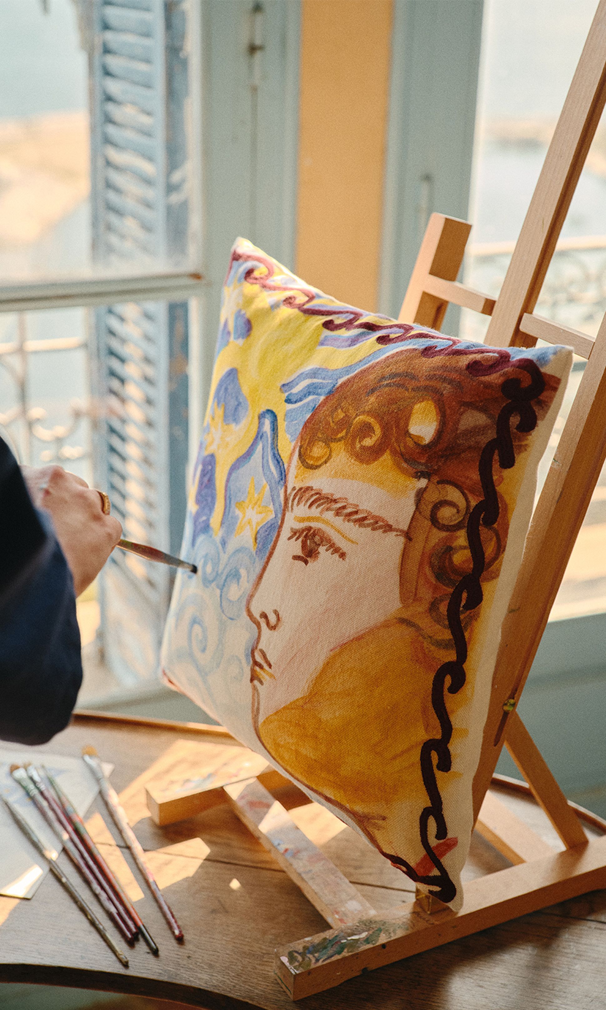 Un artiste peint un portrait haut en couleur sur un coussin posé sur un chevalet, avec des pinceaux et de la peinture à proximité, près d'une fenêtre baignée de soleil.