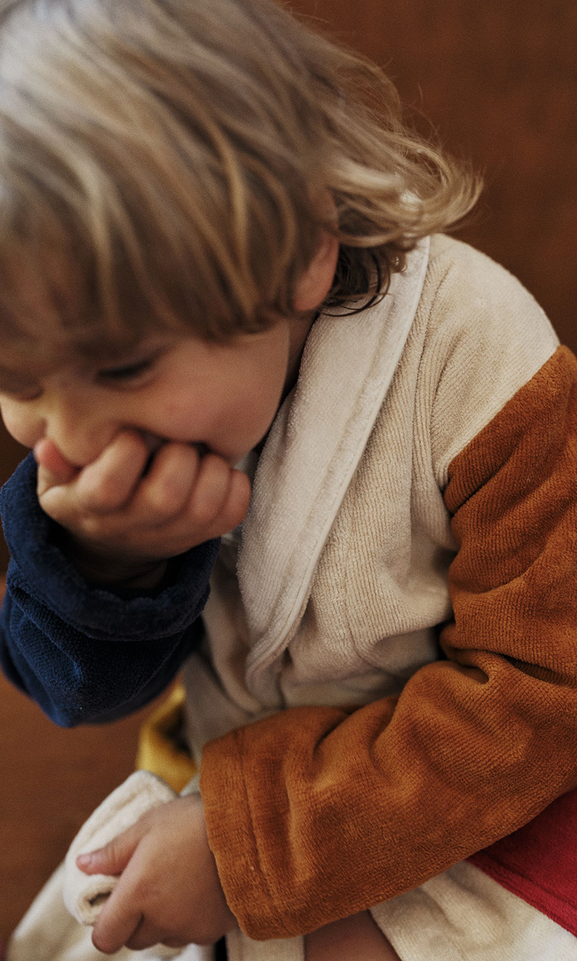 Un enfant aux cheveux blonds, vêtu d'une robe colorée, se couvre la bouche d'une main et semble joyeux dans un cadre chaleureux.