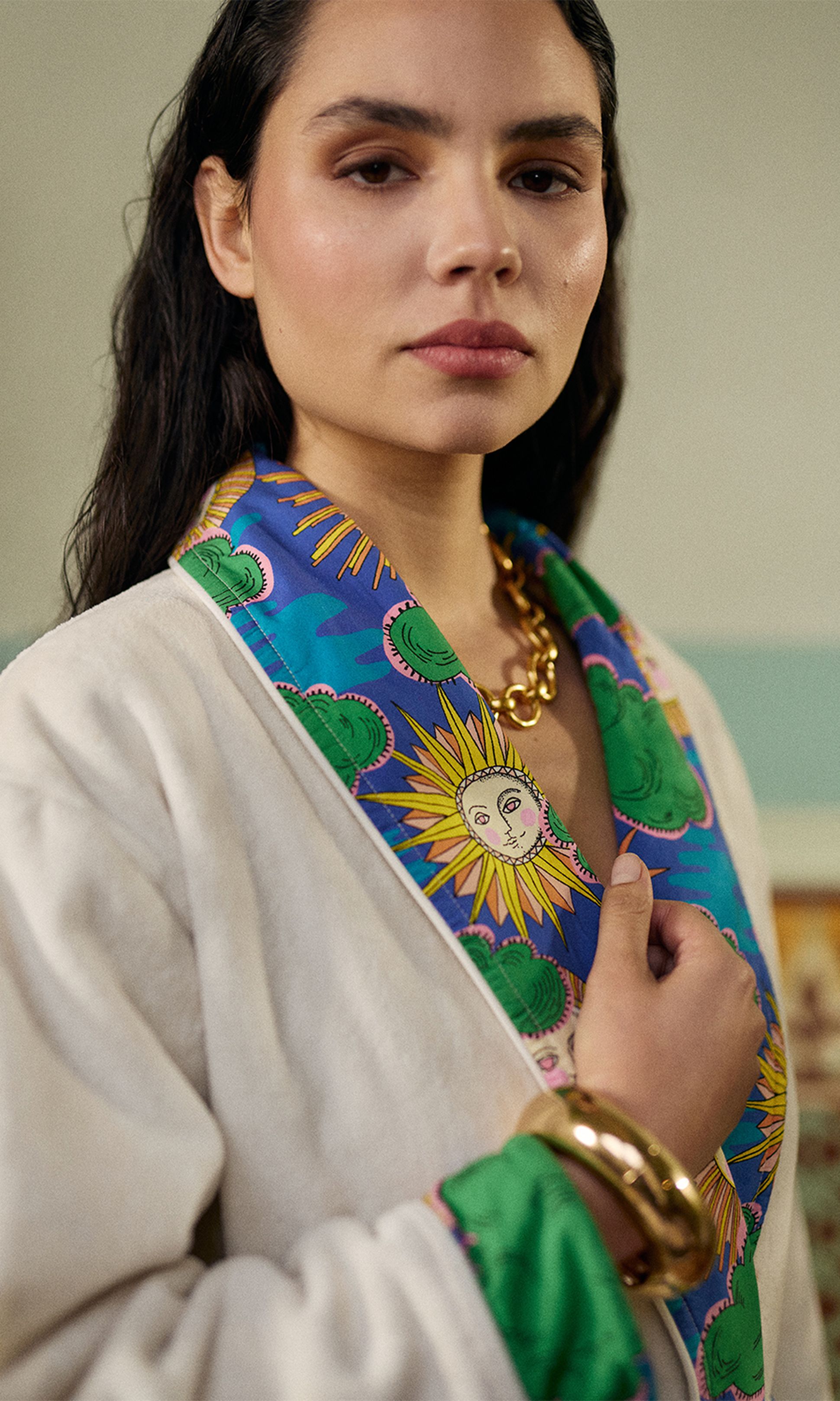 Une femme vêtue d'un peignoir blanc orné d'un col coloré à motifs de soleil, portant un gros bracelet et un collier en or, le regard assur