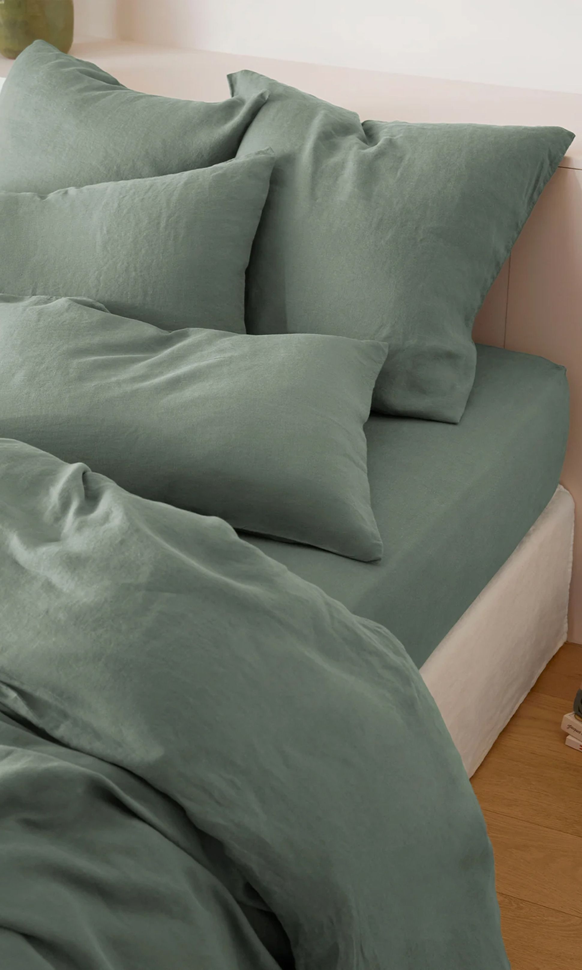 Un lit soigneusement fait, avec des oreillers et une couette en lin vert, posé sur un parquet. Une petite pile de livres est visible à côté du lit.