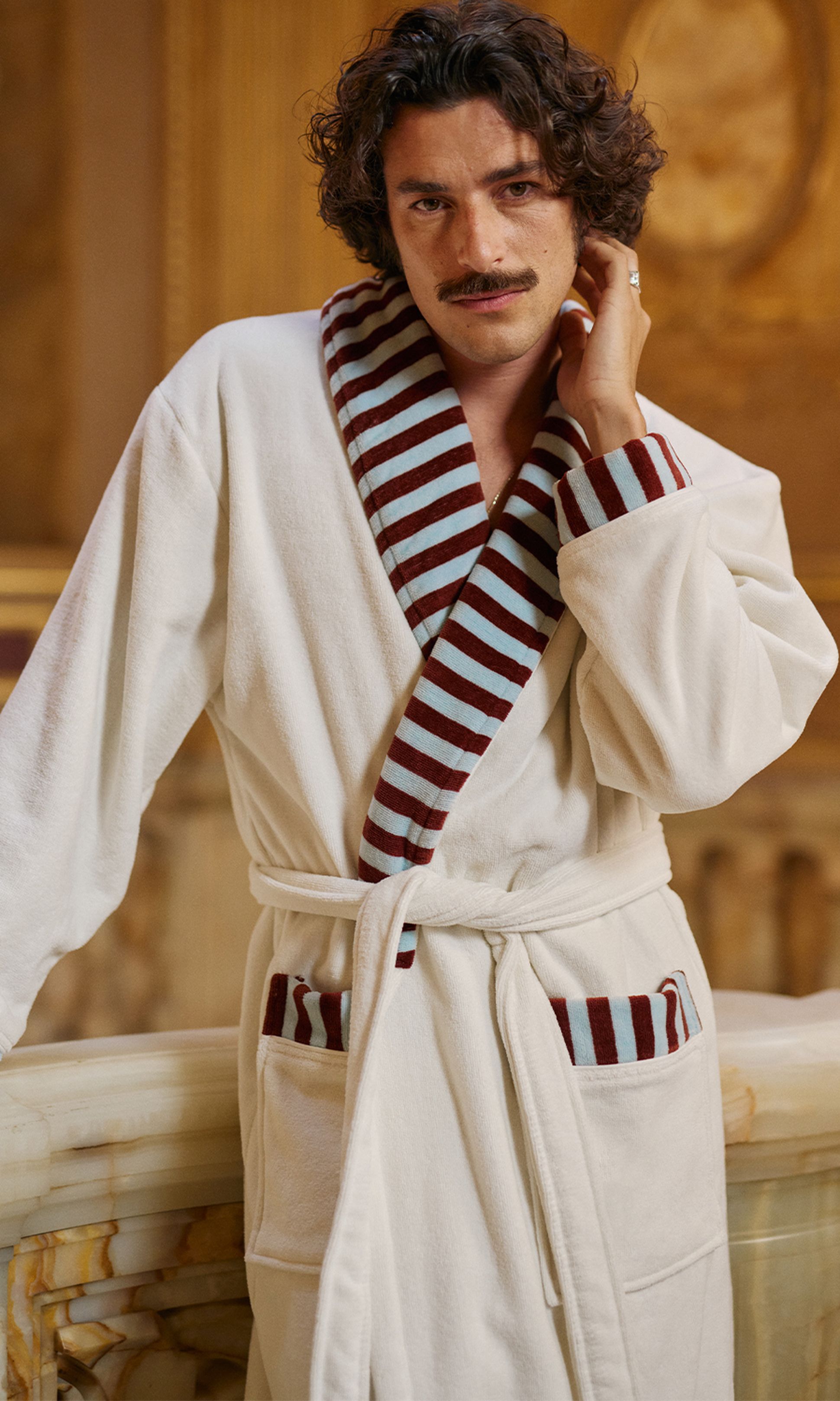 Une personne aux cheveux bouclés et portant une moustache, vêtue d'une robe blanche ornée de rayures marron et bleues, debout dans une pièce élégante.