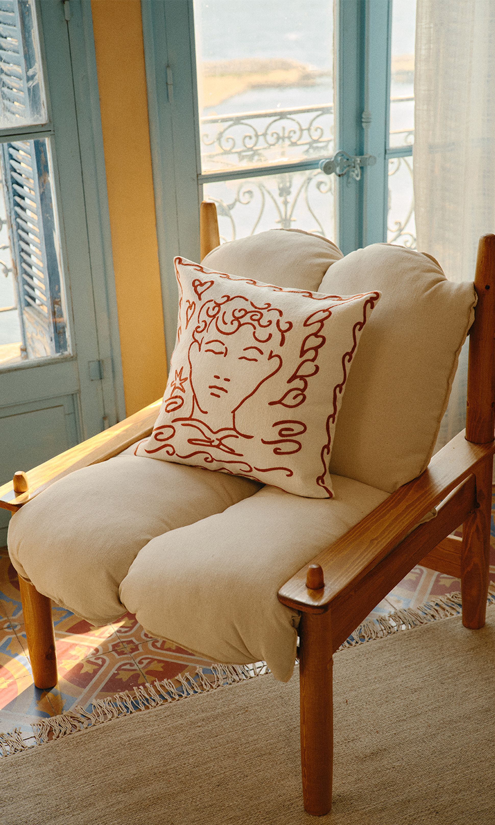 Une chaise en bois confortable, avec un coussin beige et un oreiller orné d'un motif artistique représentant un visage, près d'une fenêtre aux volets bleus offrant une vue sur la mer.
