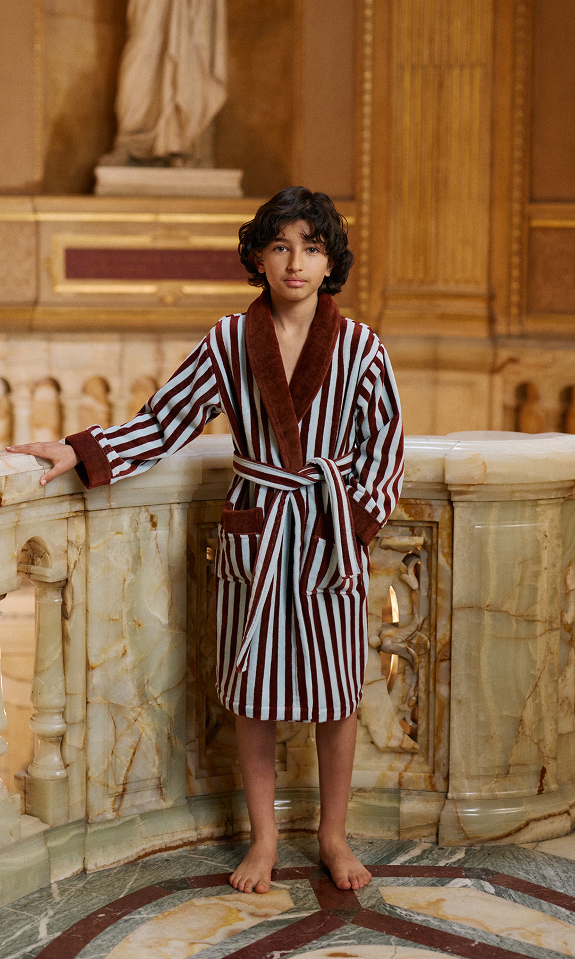 Un enfant vêtu d'une robe rayée se tient pieds nus sur un sol en marbre richement décoré, avec une statue classique en arrière-plan.