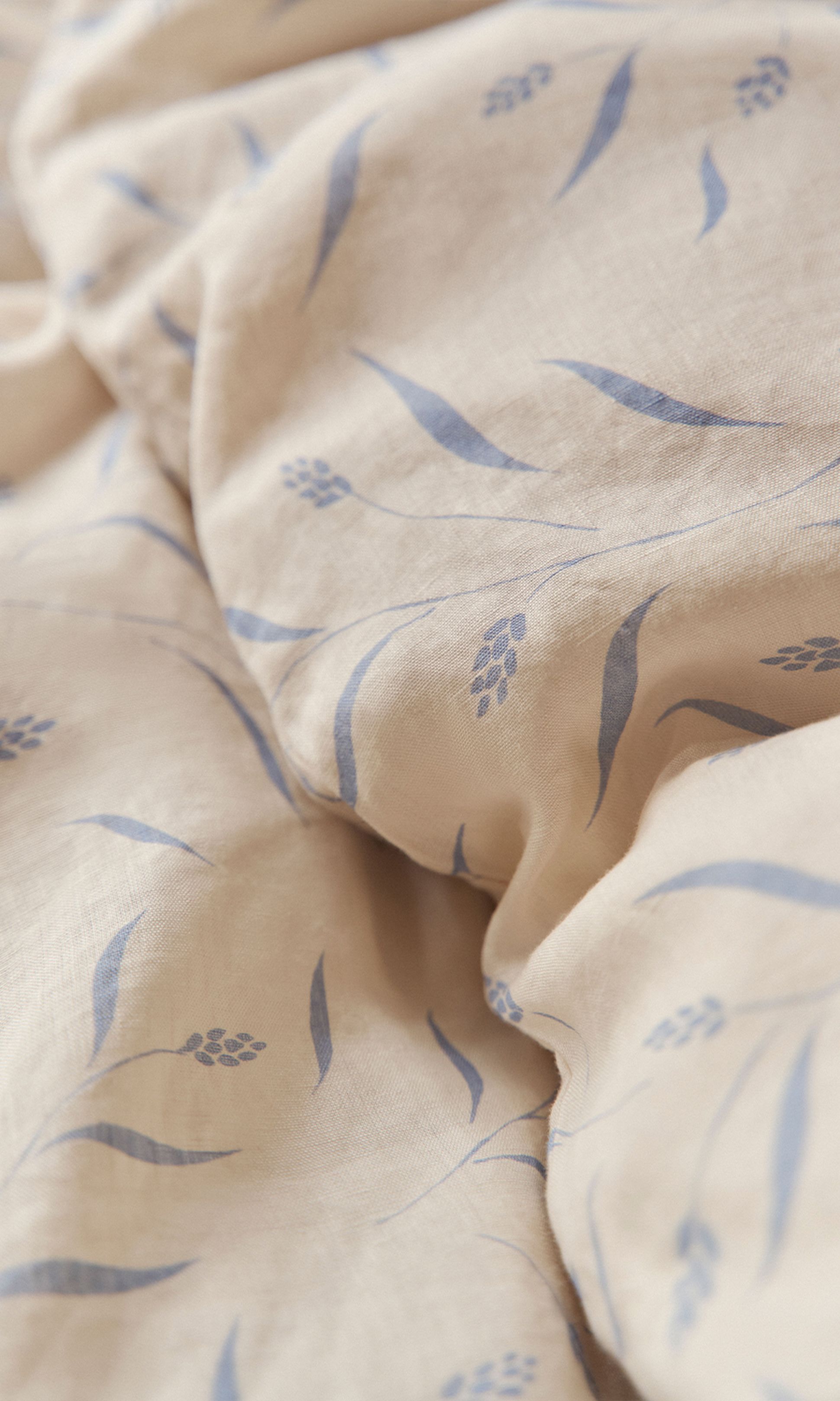 Tissu beige orné d'un motif floral bleu pâle composé de feuilles et de tiges délicates, à la texture légèrement froissée.