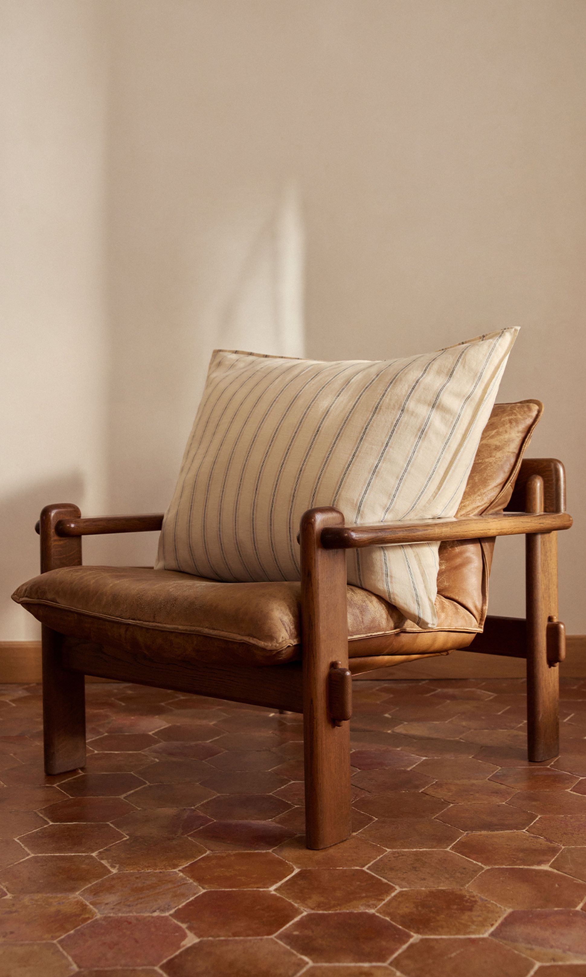 Fauteuil en bois avec des coussins en cuir marron et un grand coussin rayé, posé sur un sol carrelé d'hexagones dans une pièce baignée d'une lumière tamisée.