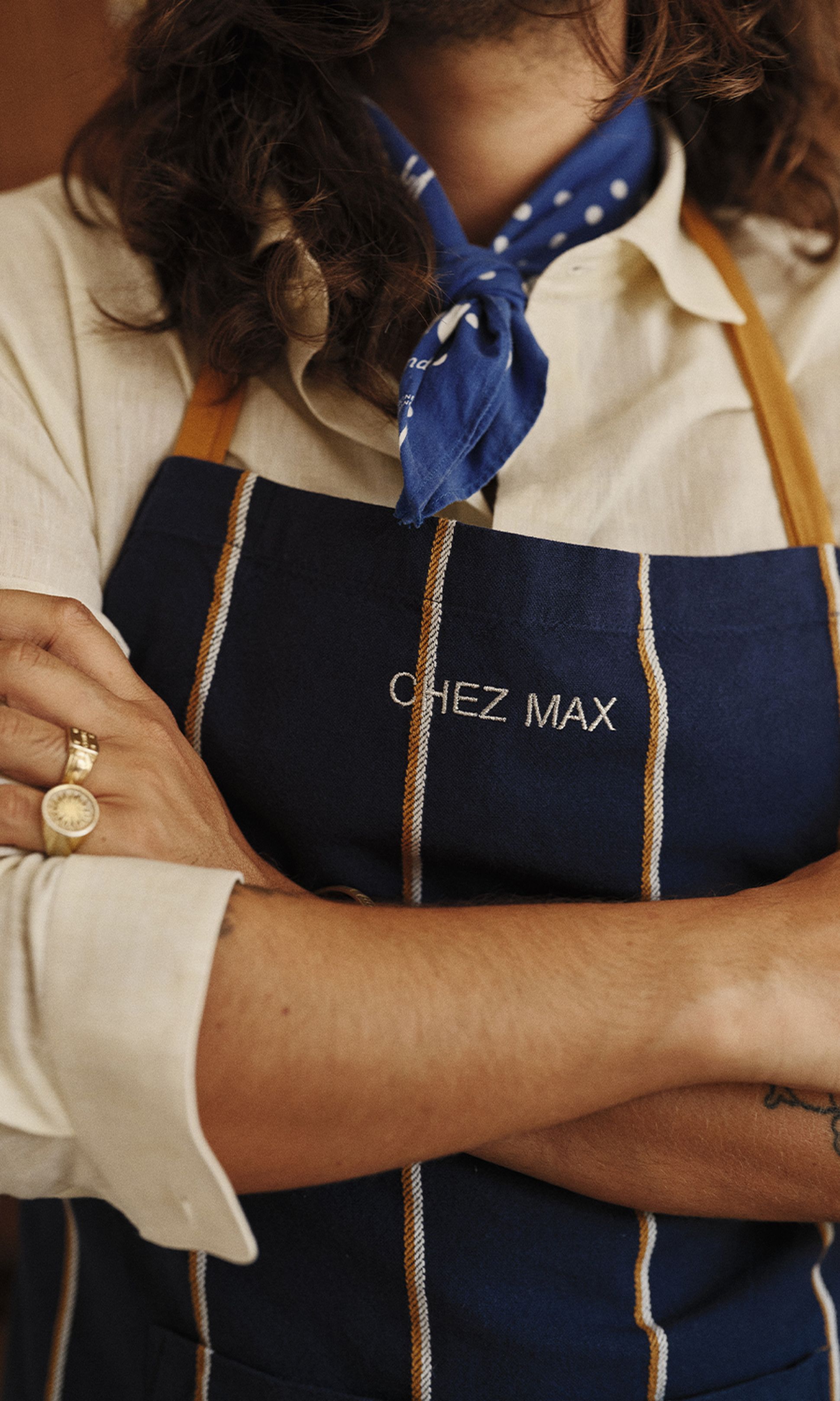 Personne aux bras croisés, portant un tablier bleu marine brodé « CHEZ MAX », une écharpe bleue et des bagues en or.