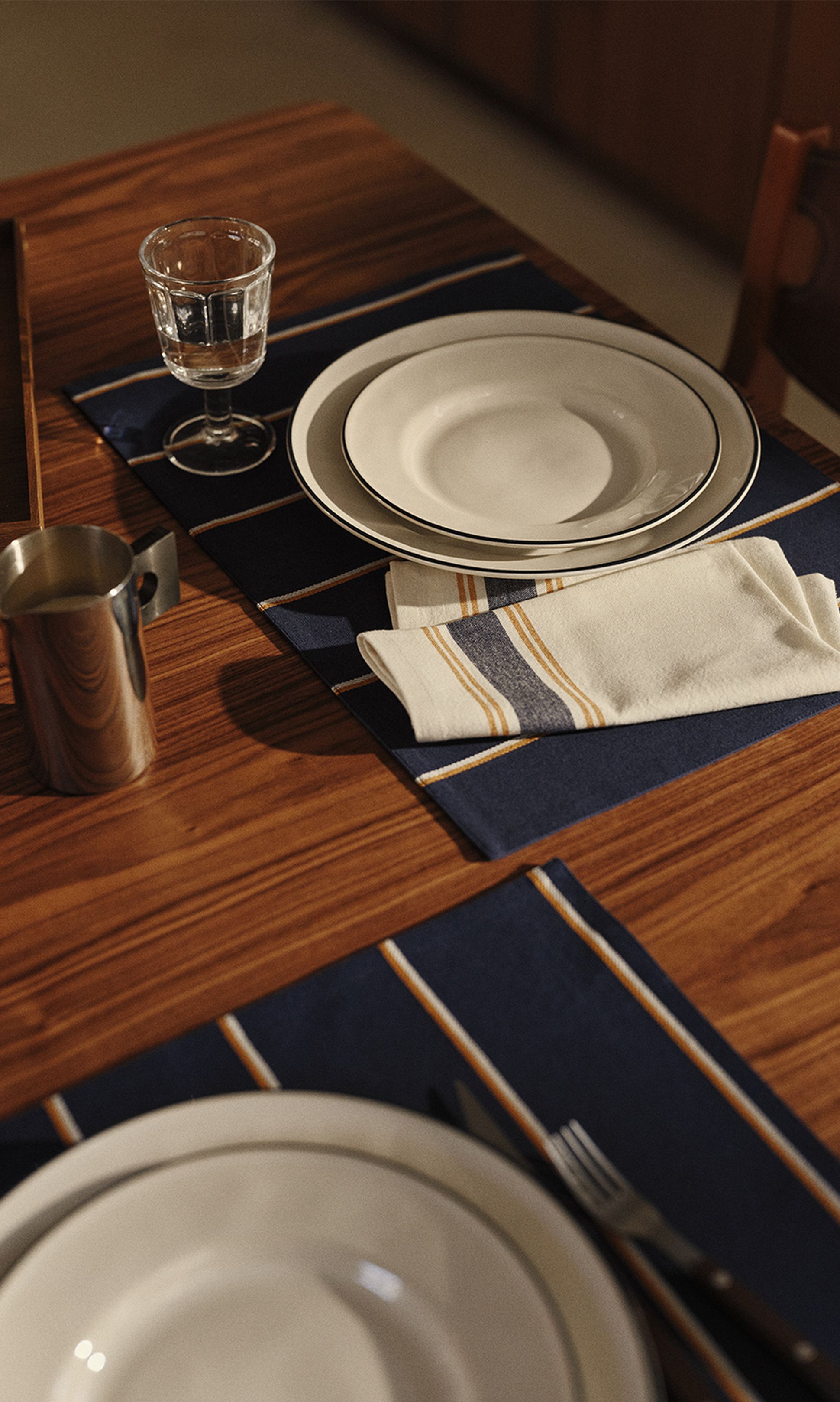 Une table élégamment dressée avec des assiettes blanches posées sur des sets de table foncés, une serviette rayée pliée, un verre à eau et une tasse en métal sur une table en bois.