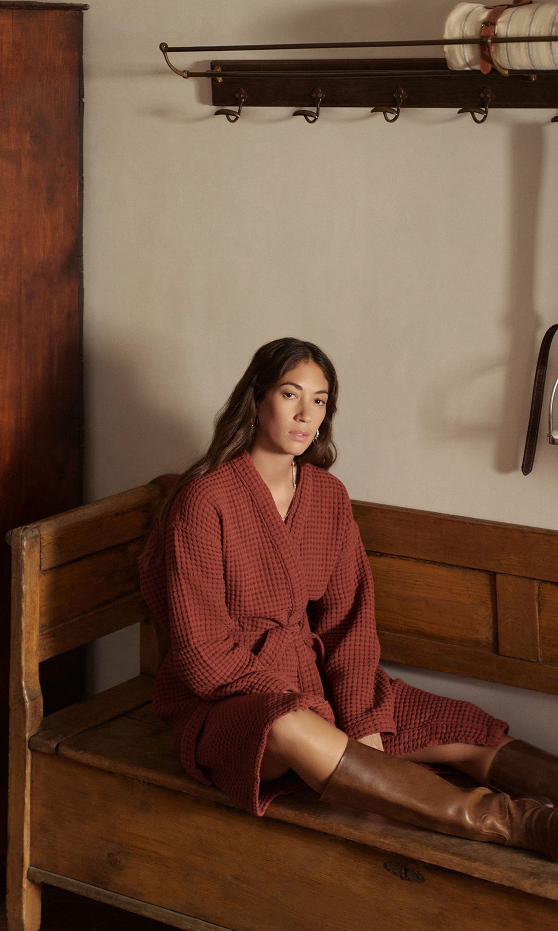 Une femme vêtue d'un peignoir rouge et de bottes marron est assise sur un banc en bois dans une pièce rustique équipée d'une armoire et d'une étagère murale sur laquelle est posée une couverture enroulée.