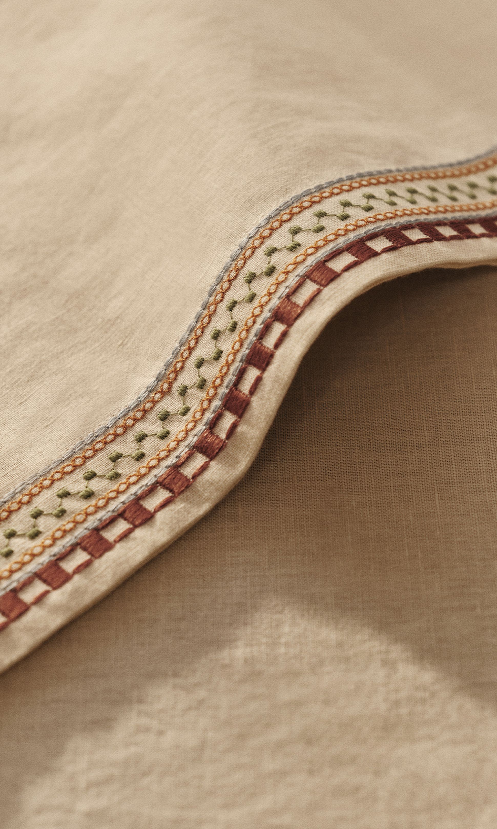 Gros plan sur un tissu beige orné de broderies détaillées en rouge, vert et orange, mettant en valeur des motifs complexes le long du bord.