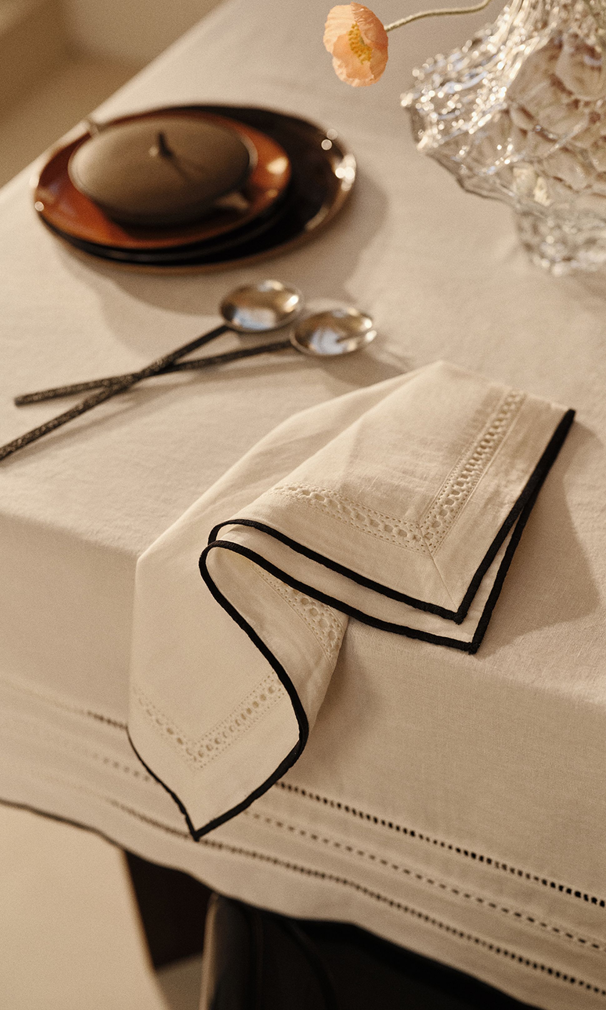 Une table élégamment dressée, avec une serviette blanche pliée aux bords noirs, deux cuillères, des assiettes empilées et un plat décoratif en verre, le tout baigné d'une lumière tamisée.