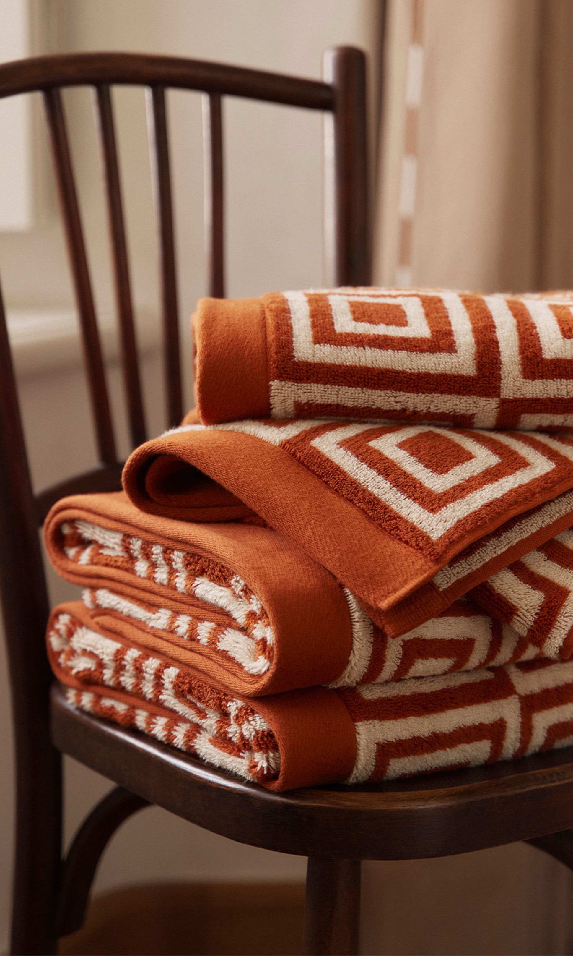 Pile de serviettes orange à motifs géométriques blancs sur une chaise en bois, placée à côté d'une fenêtre à rideaux dans une lumière tamisée.