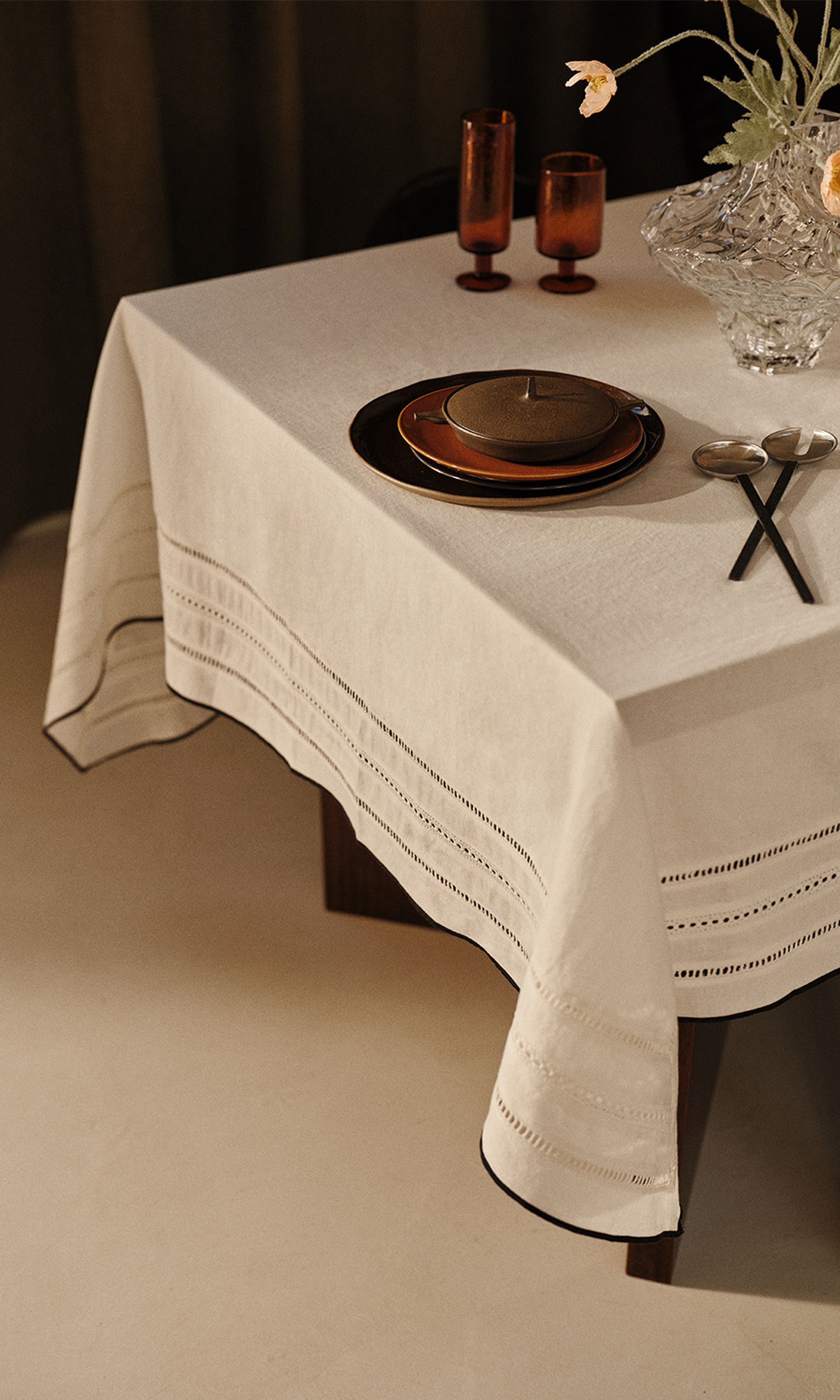 Une table élégamment dressée, avec une nappe blanche, de la vaisselle marron, un vase en cristal orné de fleurs et deux cuillères posées sur la table.