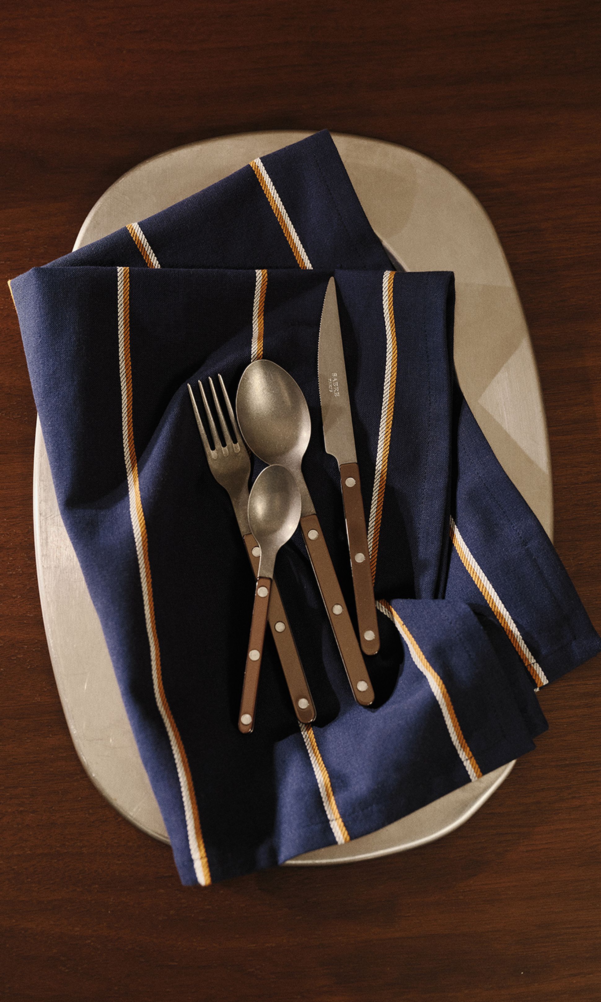 Un service de couverts aux manches marron posé sur une serviette à rayures bleues, lui-même posé sur une assiette, le tout disposé sur une table en bois.