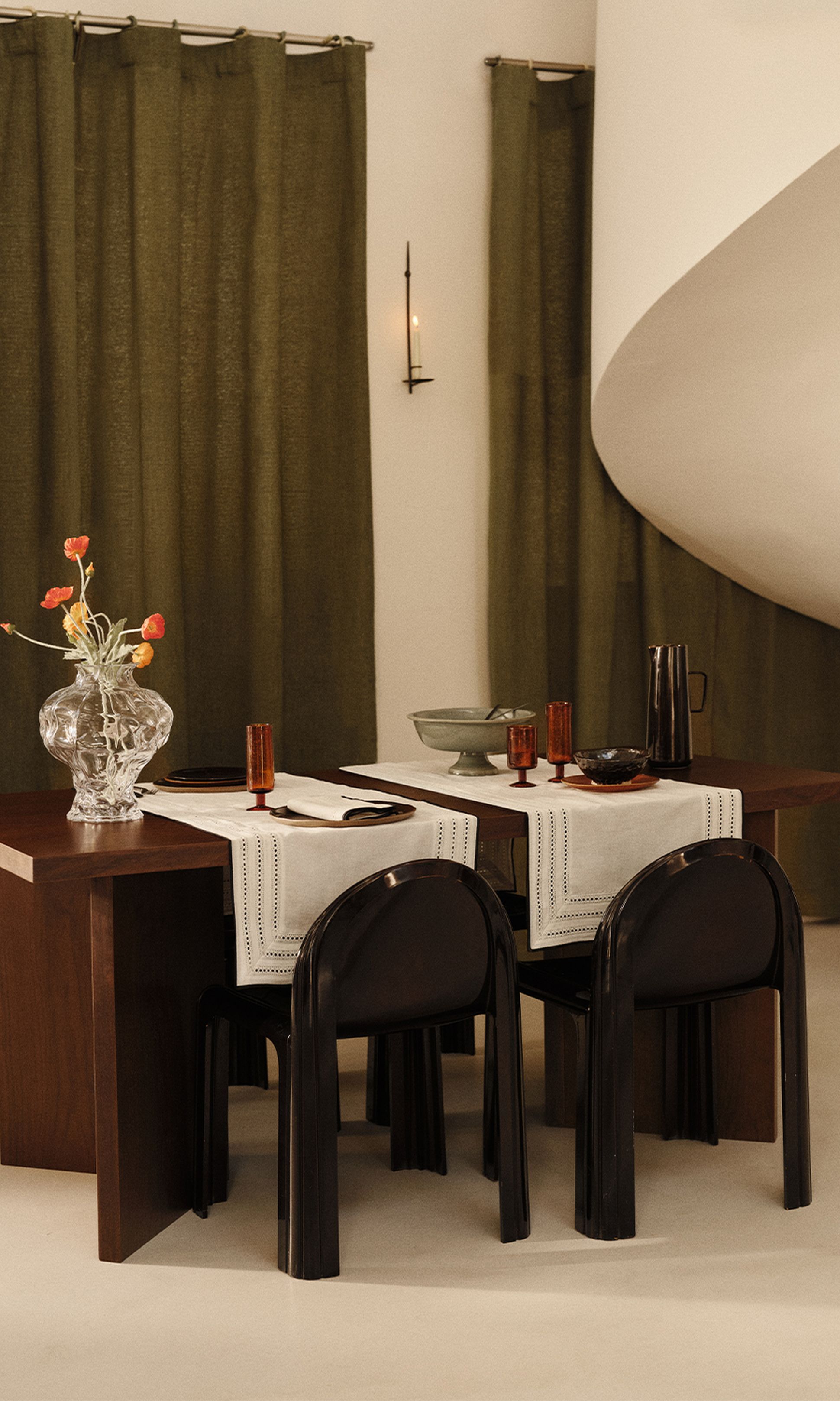 Une salle à manger élégante, avec une table en bois foncé, des chaises noires, un vase transparent rempli de fleurs et des verres ambrés posés sur des chemins de table blancs, le tout sur fond de rideaux vert olive.
