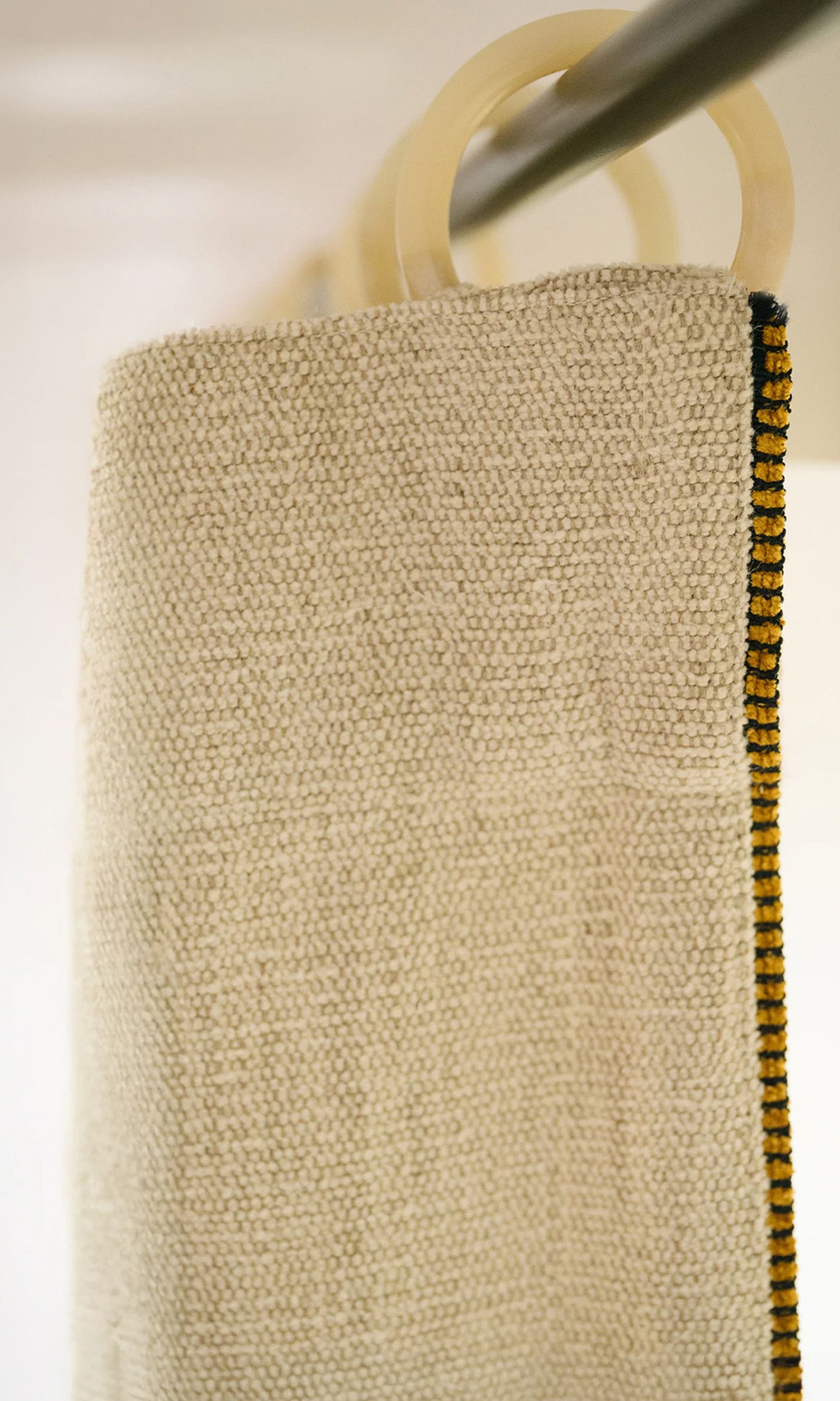 Gros plan sur un rideau beige texturé, dont l'ourlet est jaune et noir, suspendu à une tringle munie d'anneaux ronds.