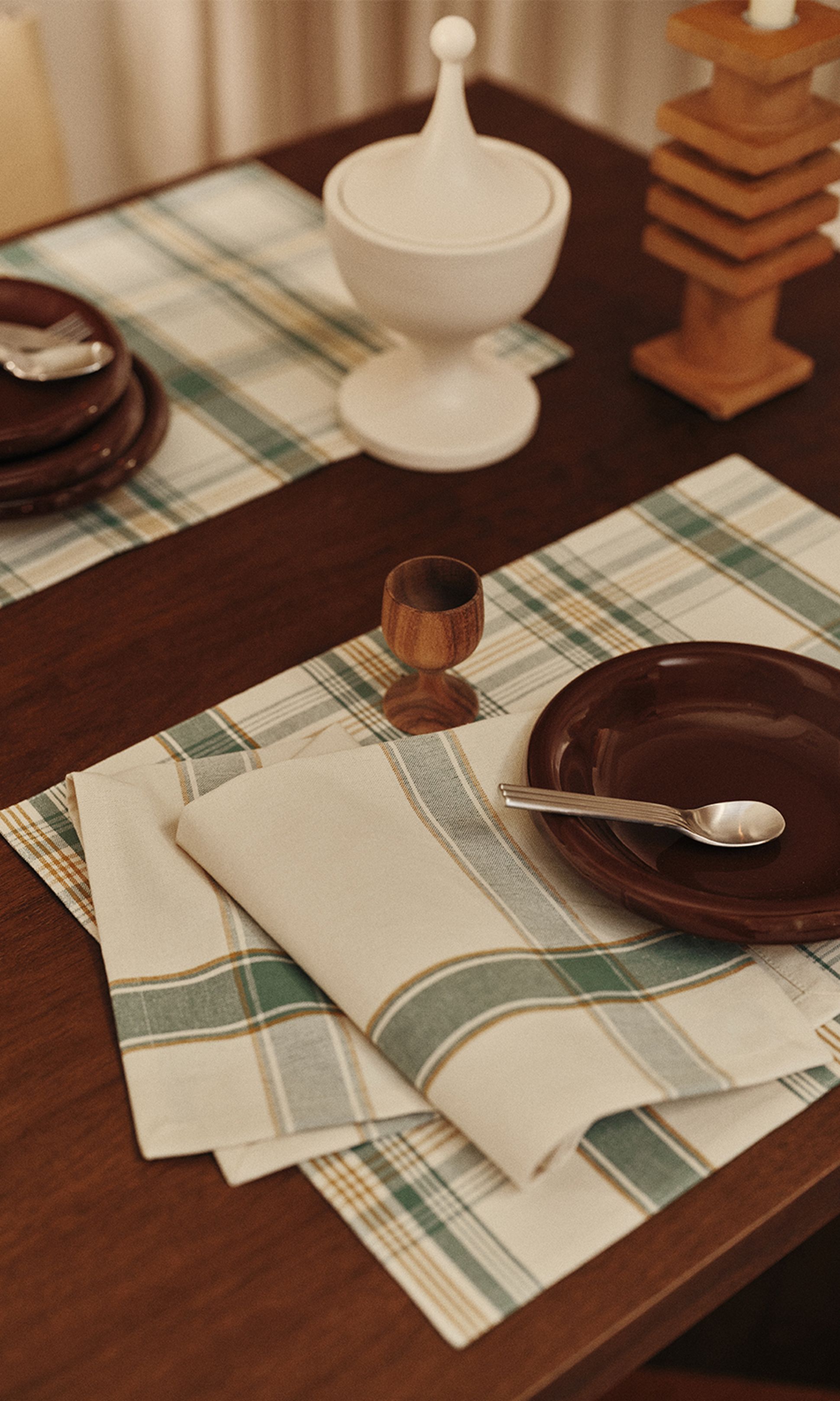 Une table dressée avec un set de table à carreaux, une assiette marron, une cuillère, une serviette pliée, un coquetier en bois et un bol muni d'un couvercle, dans une salle à manger aux tons chauds.