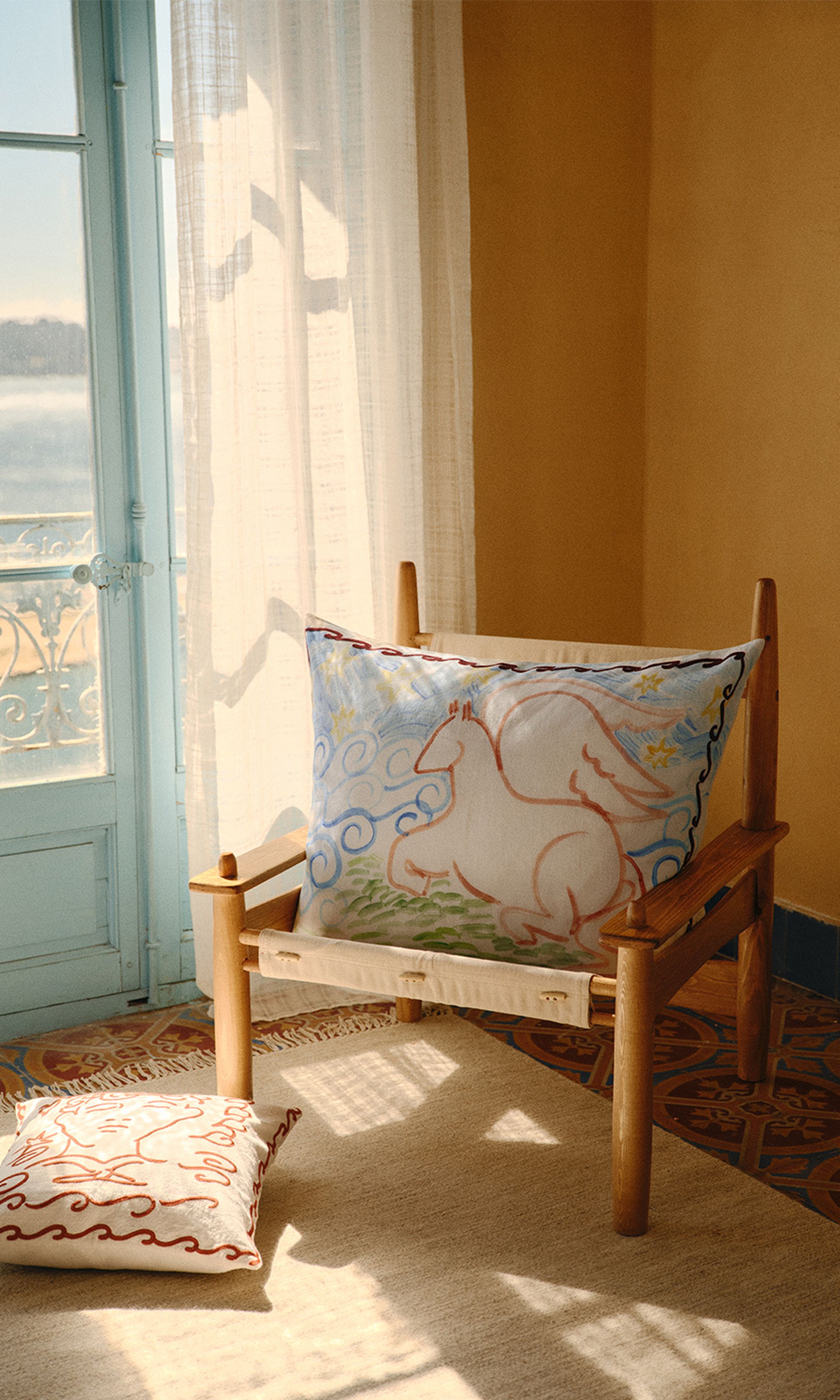 Une chaise en bois, ornée de coussins décorés d'un motif de cheval, est placée près d'une fenêtre bleue dans une pièce baignée de soleil aux murs jaunes.