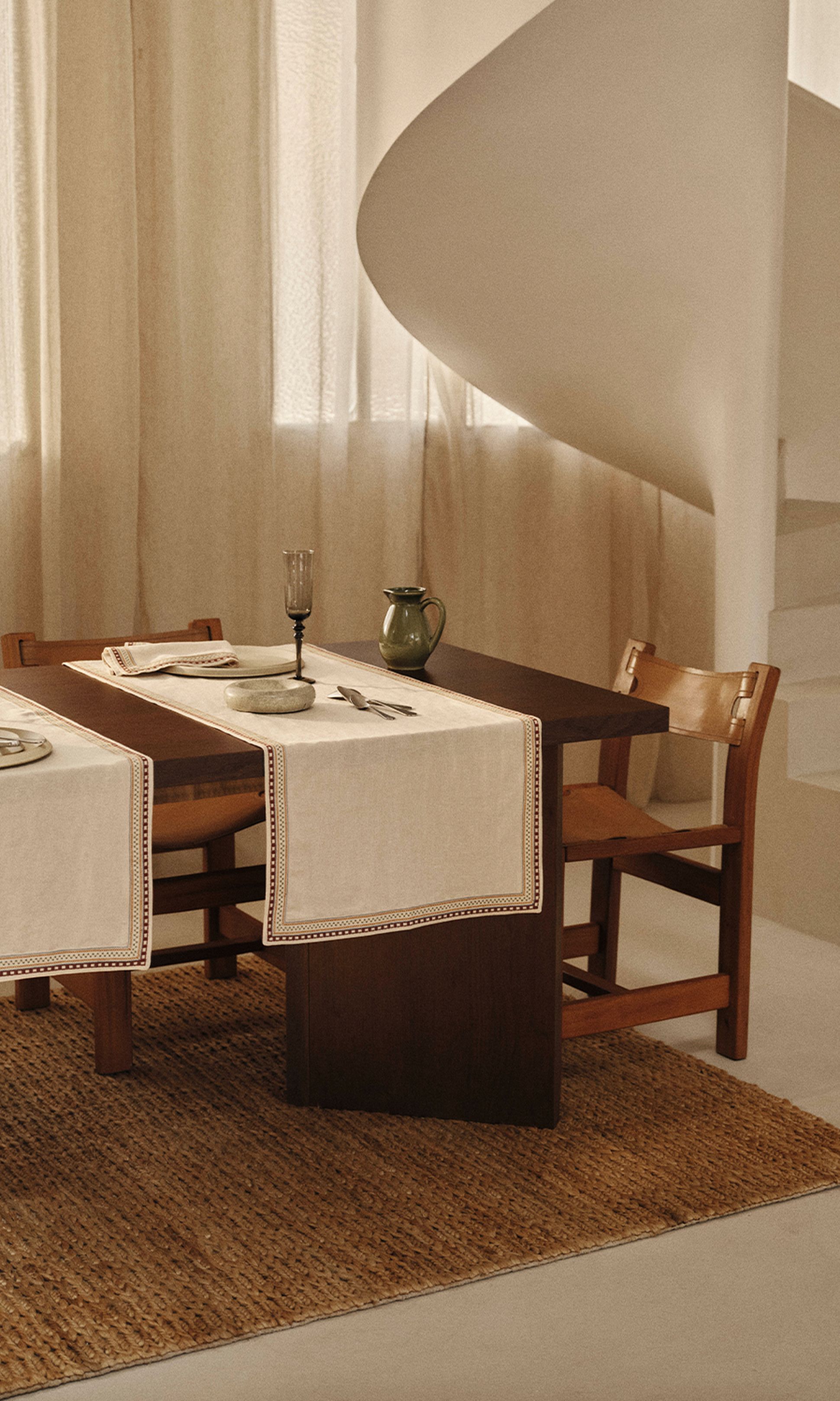 Une table à manger en bois avec des sets de table blancs, un verre et un pichet. Elle se trouve dans une pièce baignée de lumière naturelle, avec un escalier en colimaçon en arrière-plan.