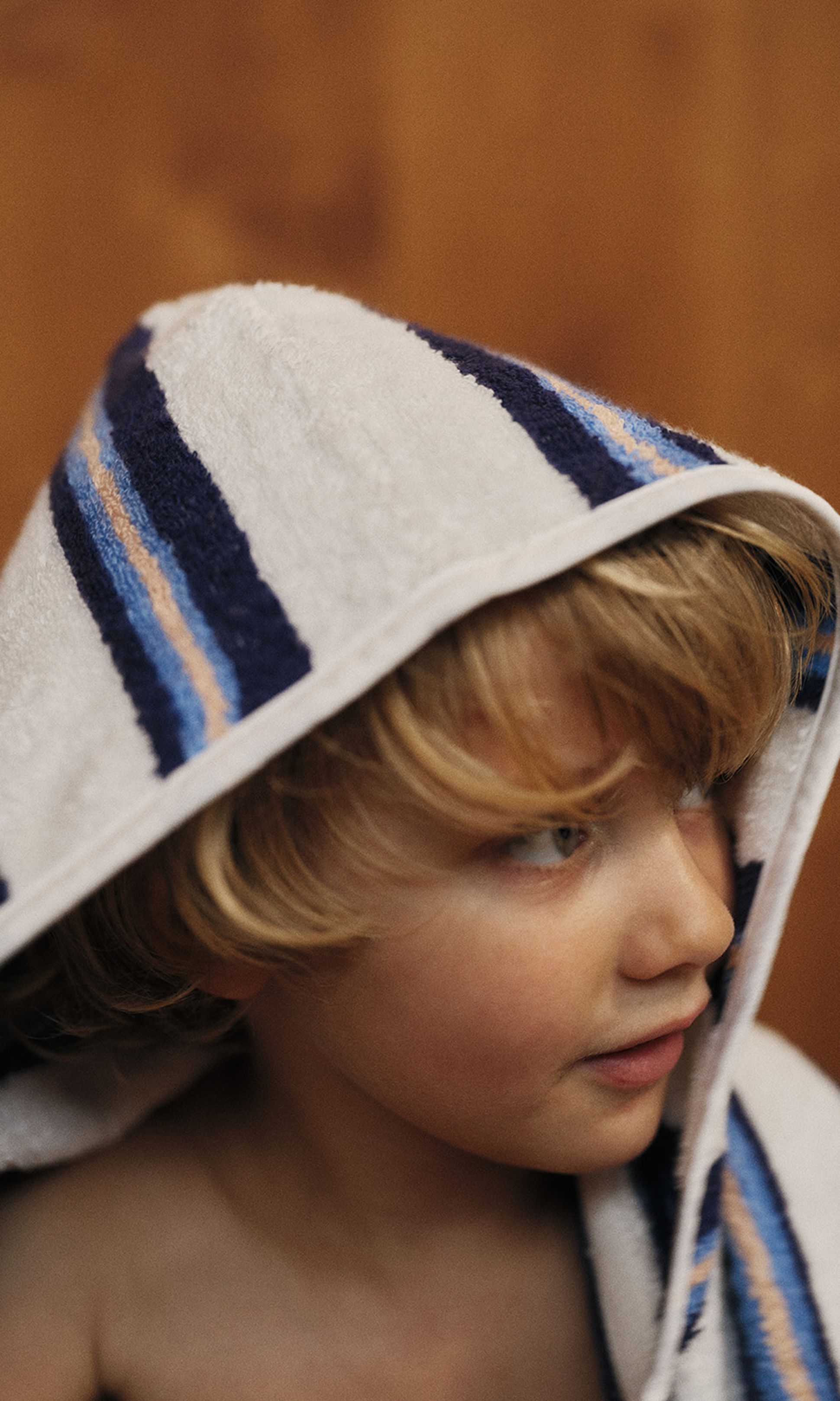 Jeune enfant aux cheveux blonds, enveloppé dans une serviette à rayures bleues, regardant sur le côté, sur un fond marron flou.