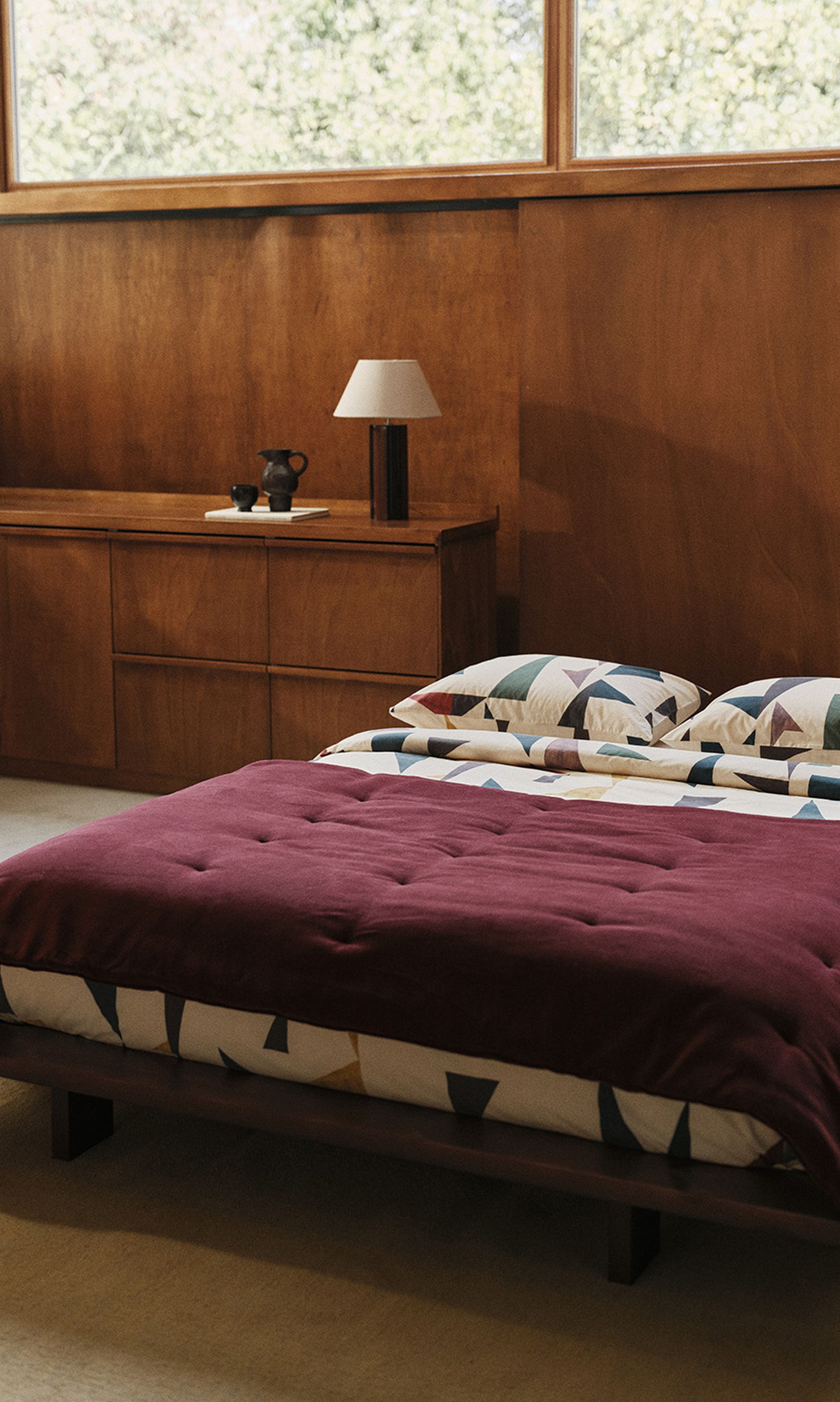 Une chambre en bois avec une couverture bordeaux sur le lit, des coussins aux motifs colorés, des commodes en bois, une lampe et de grandes fenêtres donnant sur la verdure.