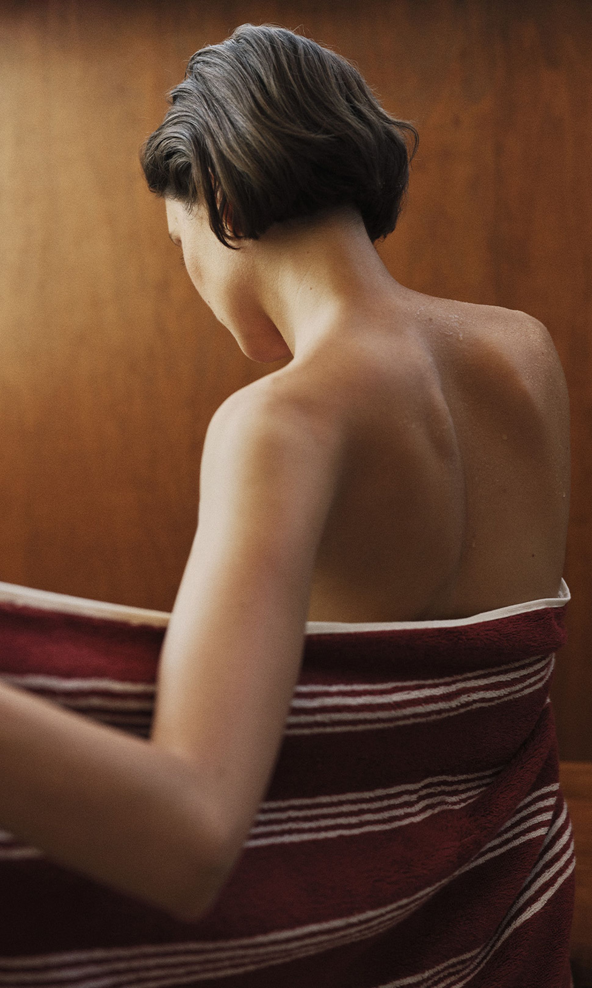 Personne aux cheveux courts, enveloppée dans une serviette rayée rouge, vue de dos sur un fond en bois.
