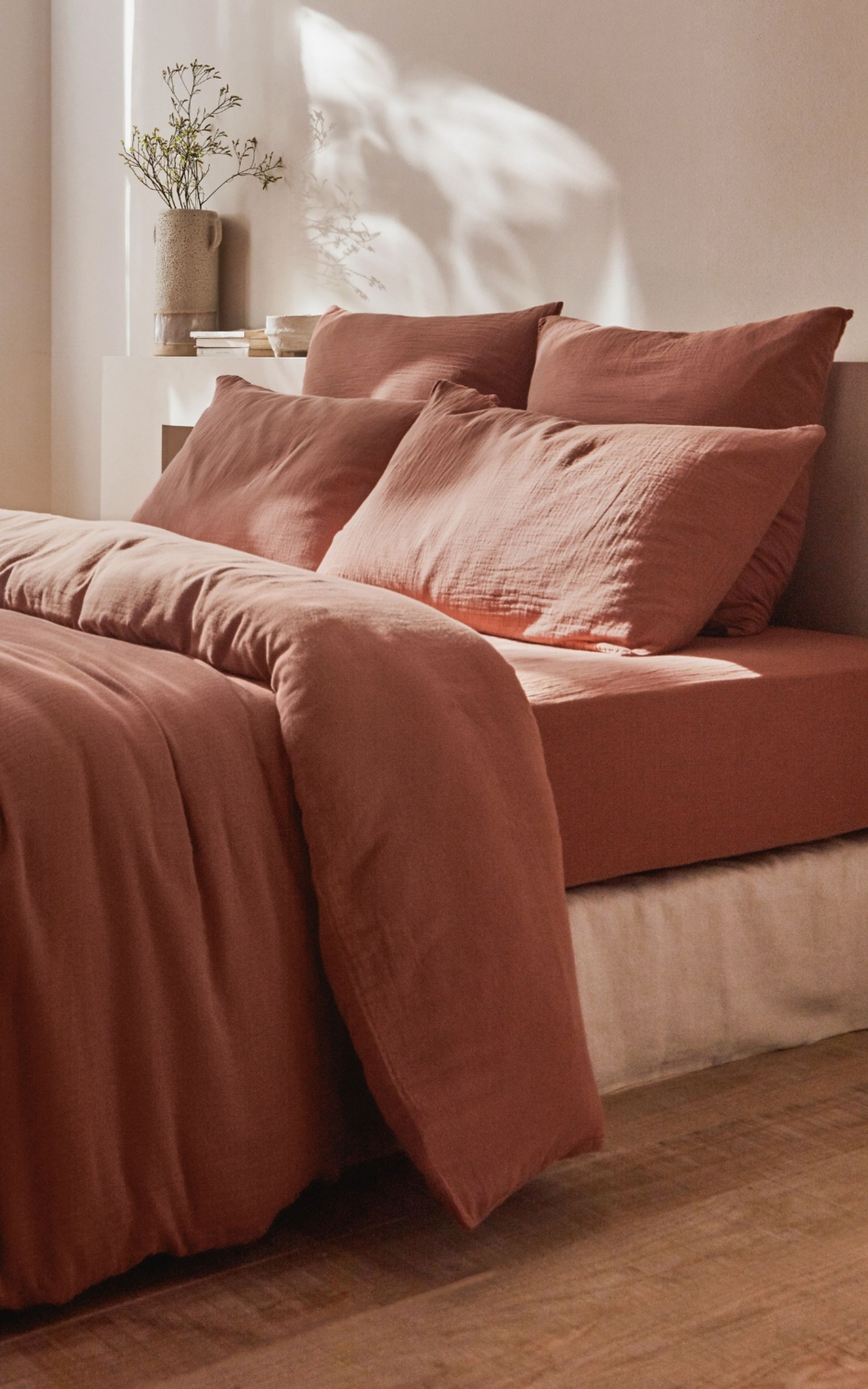 Un lit douillet avec une literie couleur rouille, adossé à un mur beige avec des étagères encastrées, et un vase avec des branches dessus.