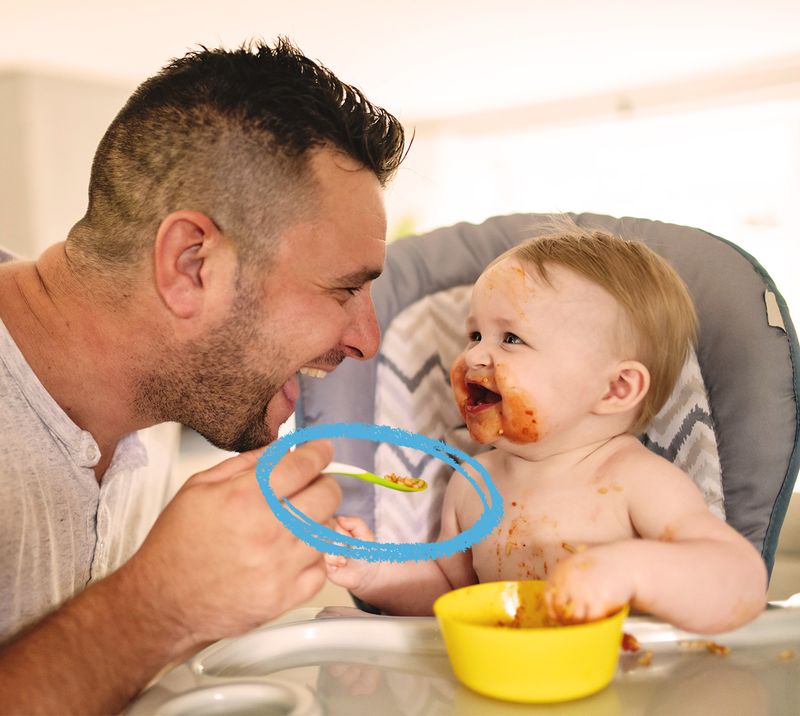 Immagine correlata al testo bambino seduto sul seggiolone che viene imboccato dal papà, entrambi sorridono