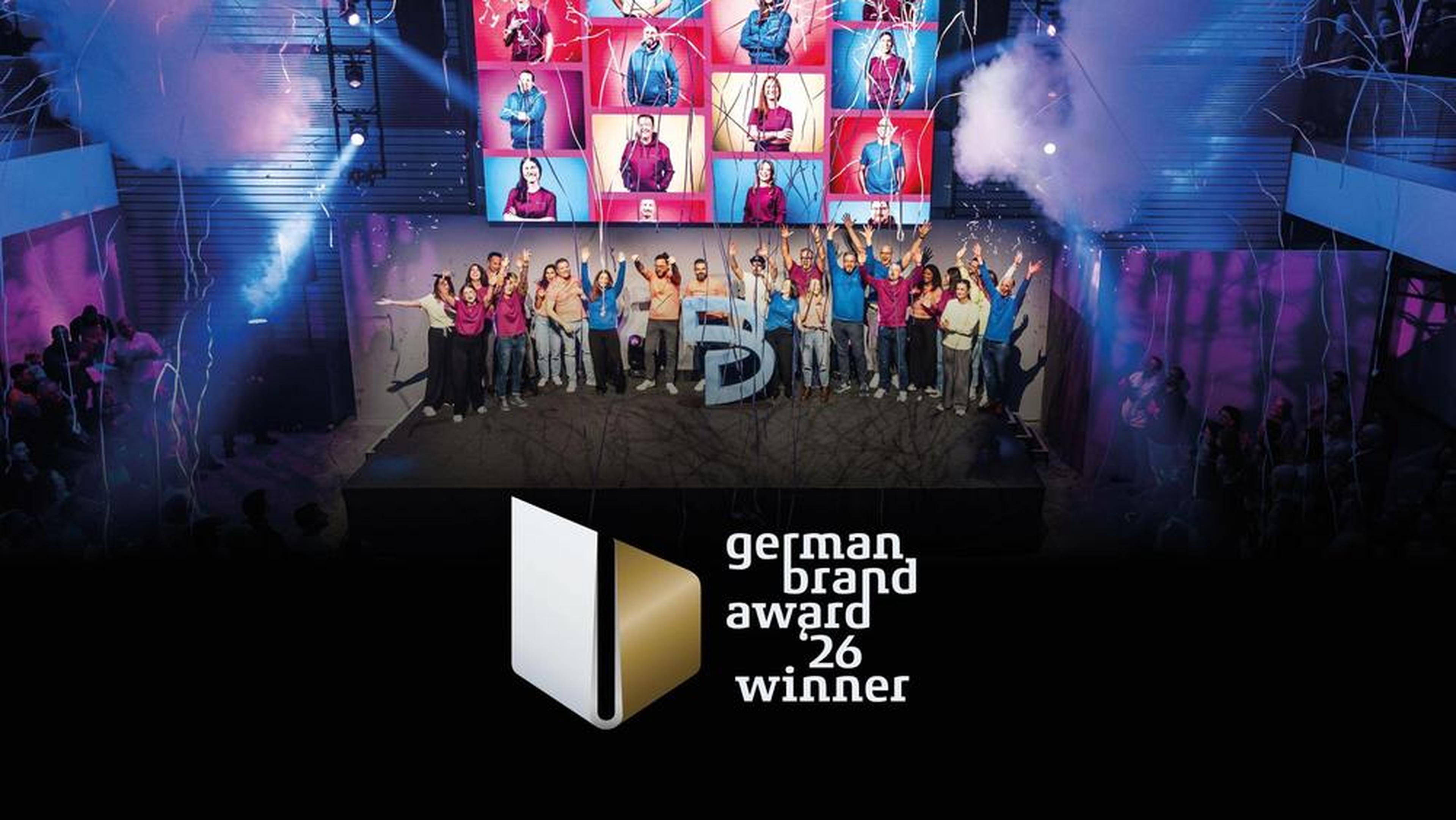People celebrating on stage with confetti and smoke, under a video wall displaying various portraits. Text reads "German Brand Award 26 Winner."