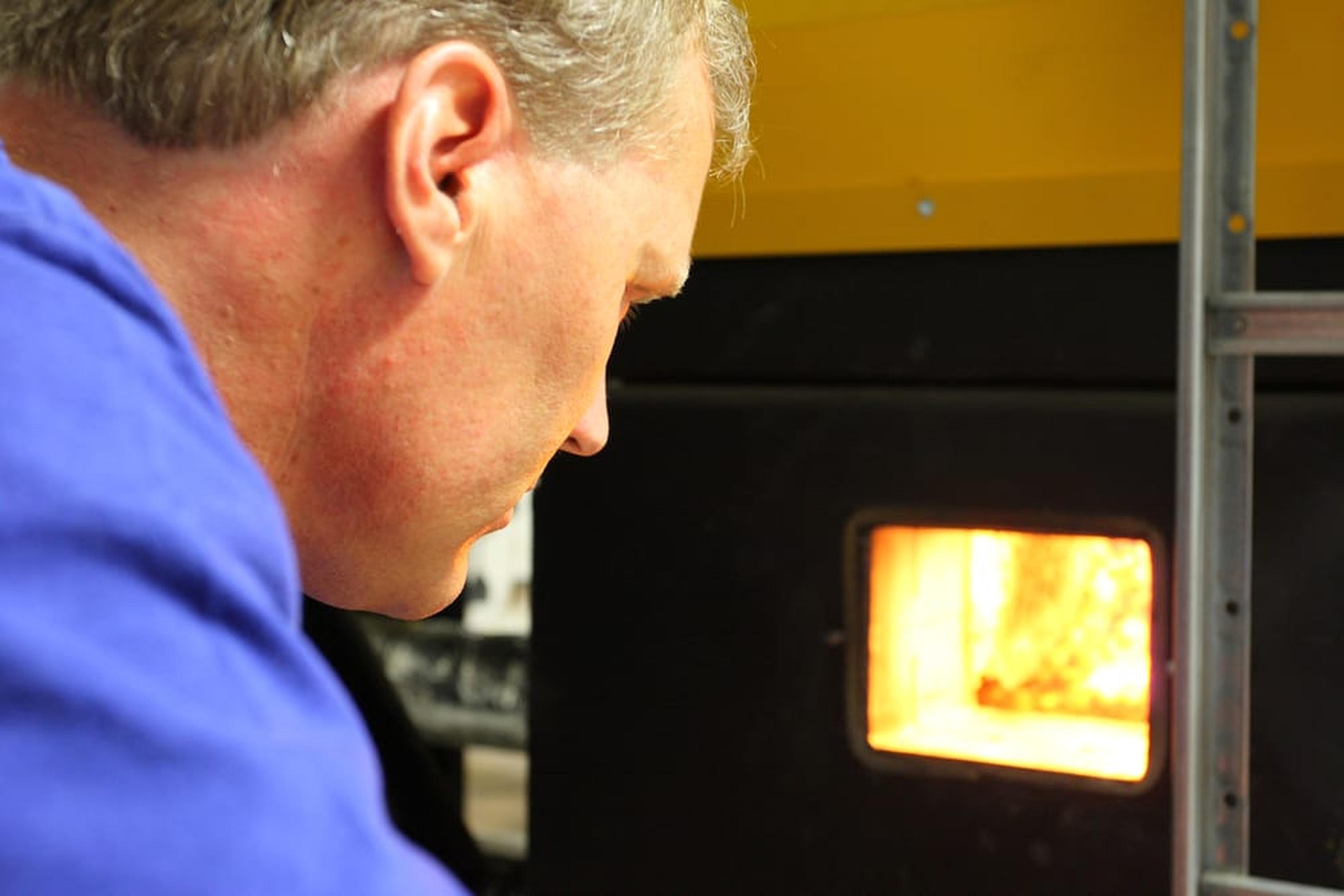 Mann in blauem Hemd beobachtet einen kleinen Ofen mit offener Tür, aus dem helle Flammen zu sehen sind.