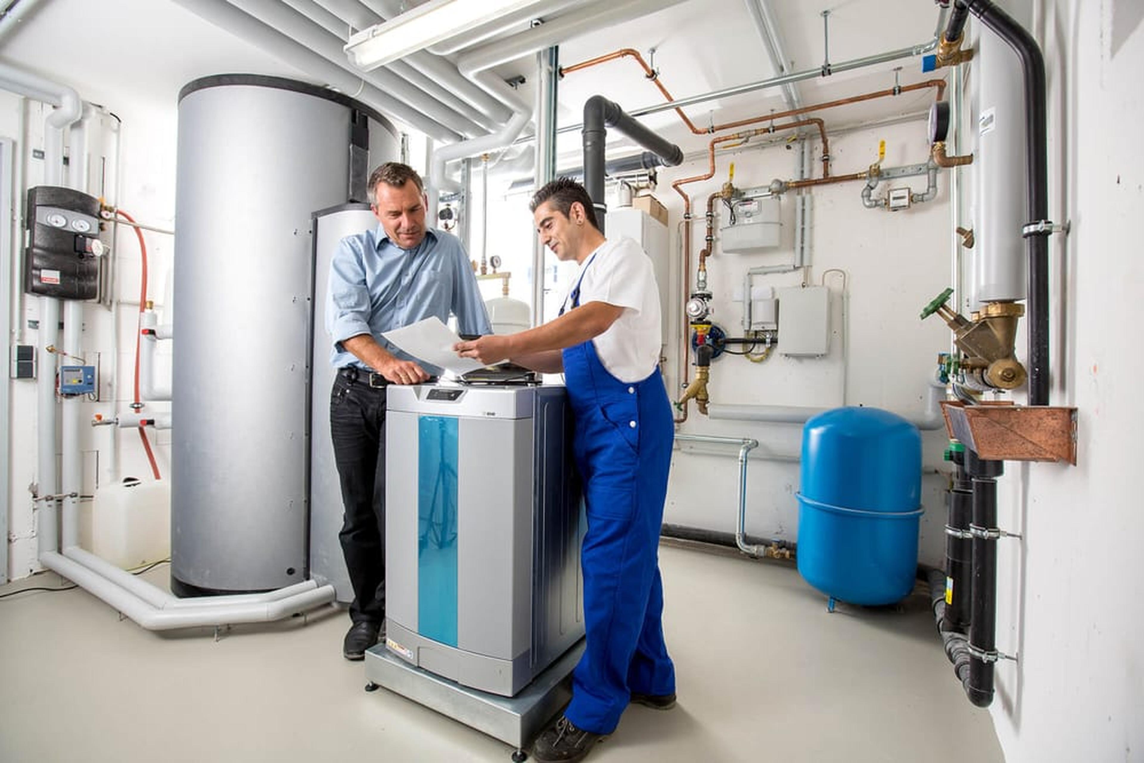 Zwei Techniker unterhalten sich in einem Hauswirtschaftsraum über eine Heizungsanlage. Der eine trägt ein blaues Hemd, der andere einen blauen Overall. Rohre und ein Tank sind zu sehen.