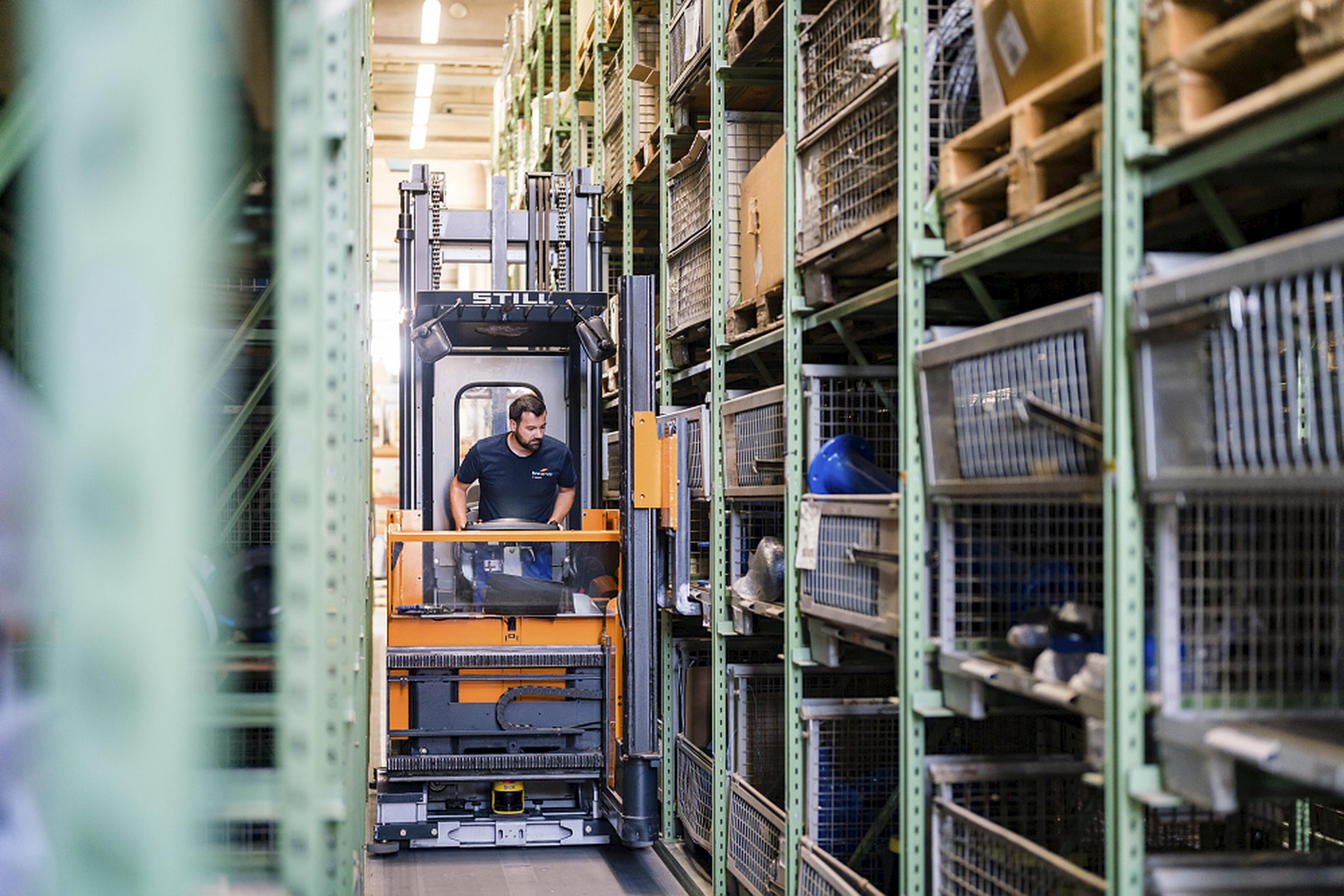 A worker operates a forklift in a warehouse aisle, surrounded by metal shelves filled with various items and containers.