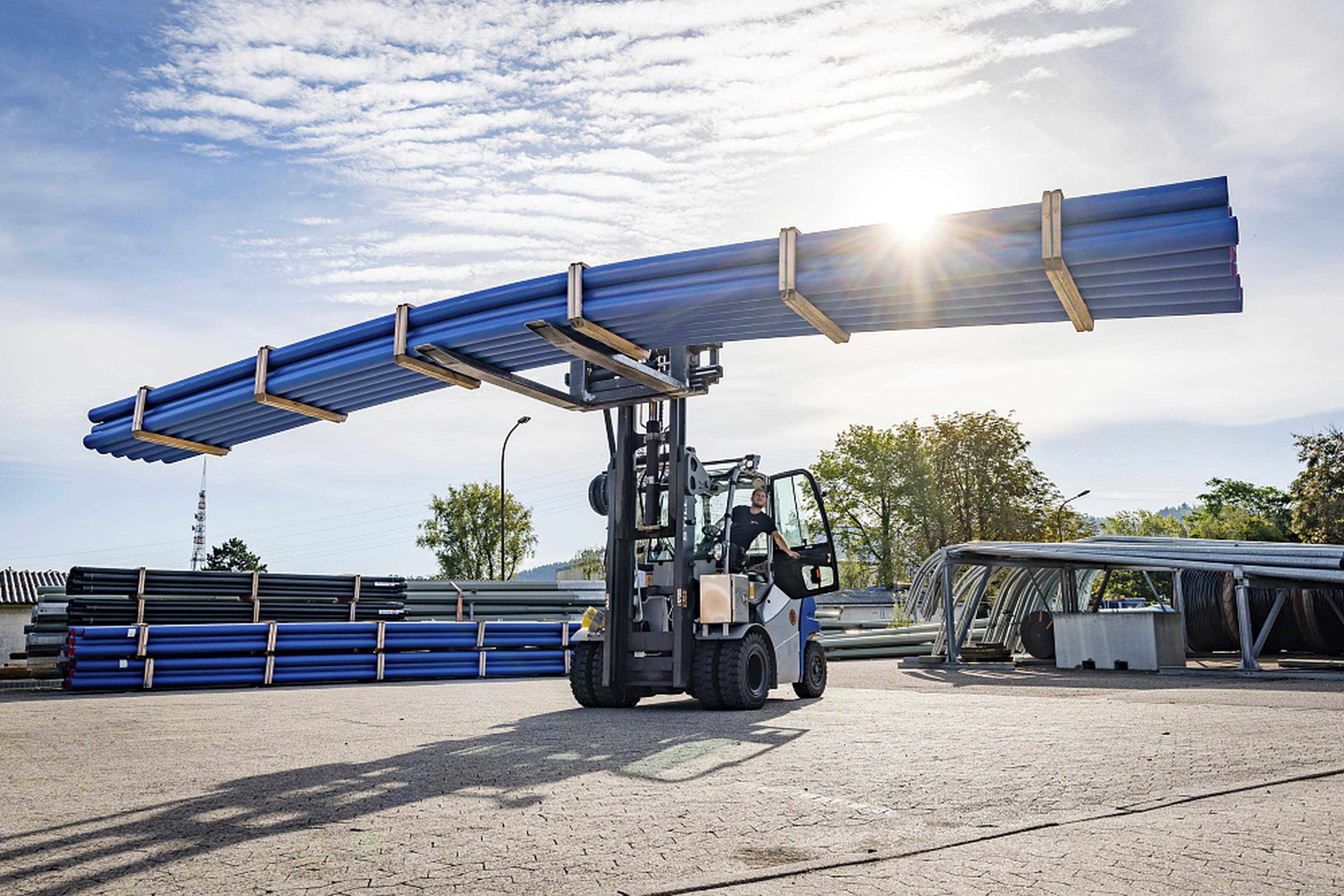 A forklift carries long blue pipes outdoors, with the sun shining through trees and stacks of pipes in the background.