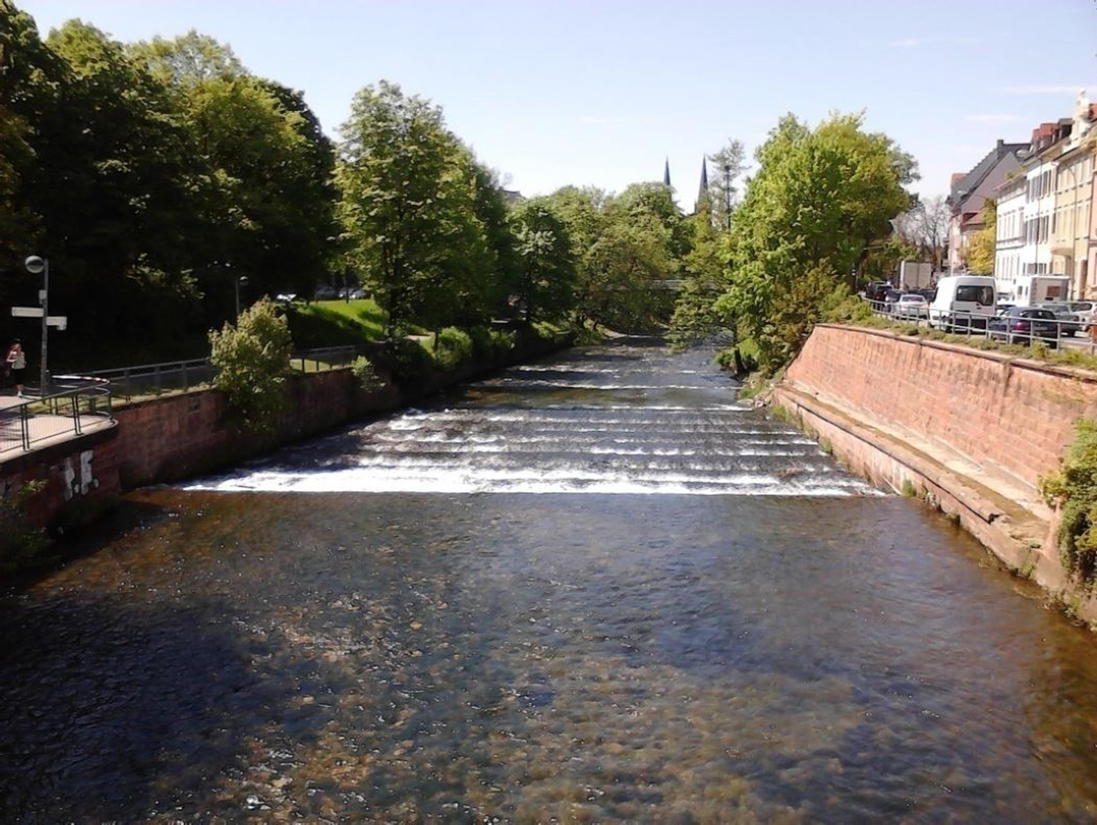 Unter einem strahlend blauen Himmel fließt ein ruhiger Fluss mit kleinen Kaskaden zwischen von Bäumen gesäumten Ufern und angrenzenden Gebäuden.