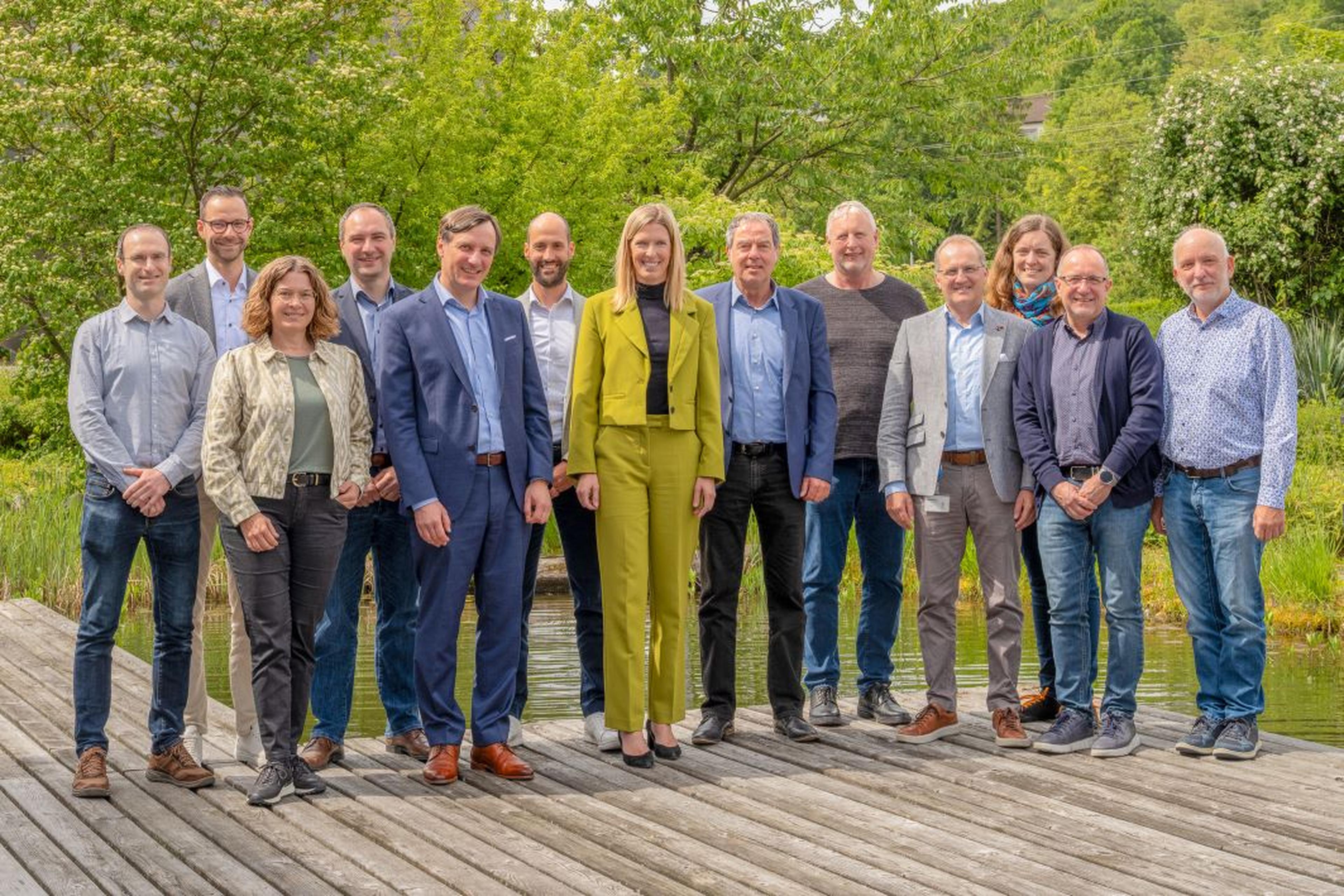 A group of thirteen people stand on a wooden deck in front of lush greenery, posing and smiling outdoors.