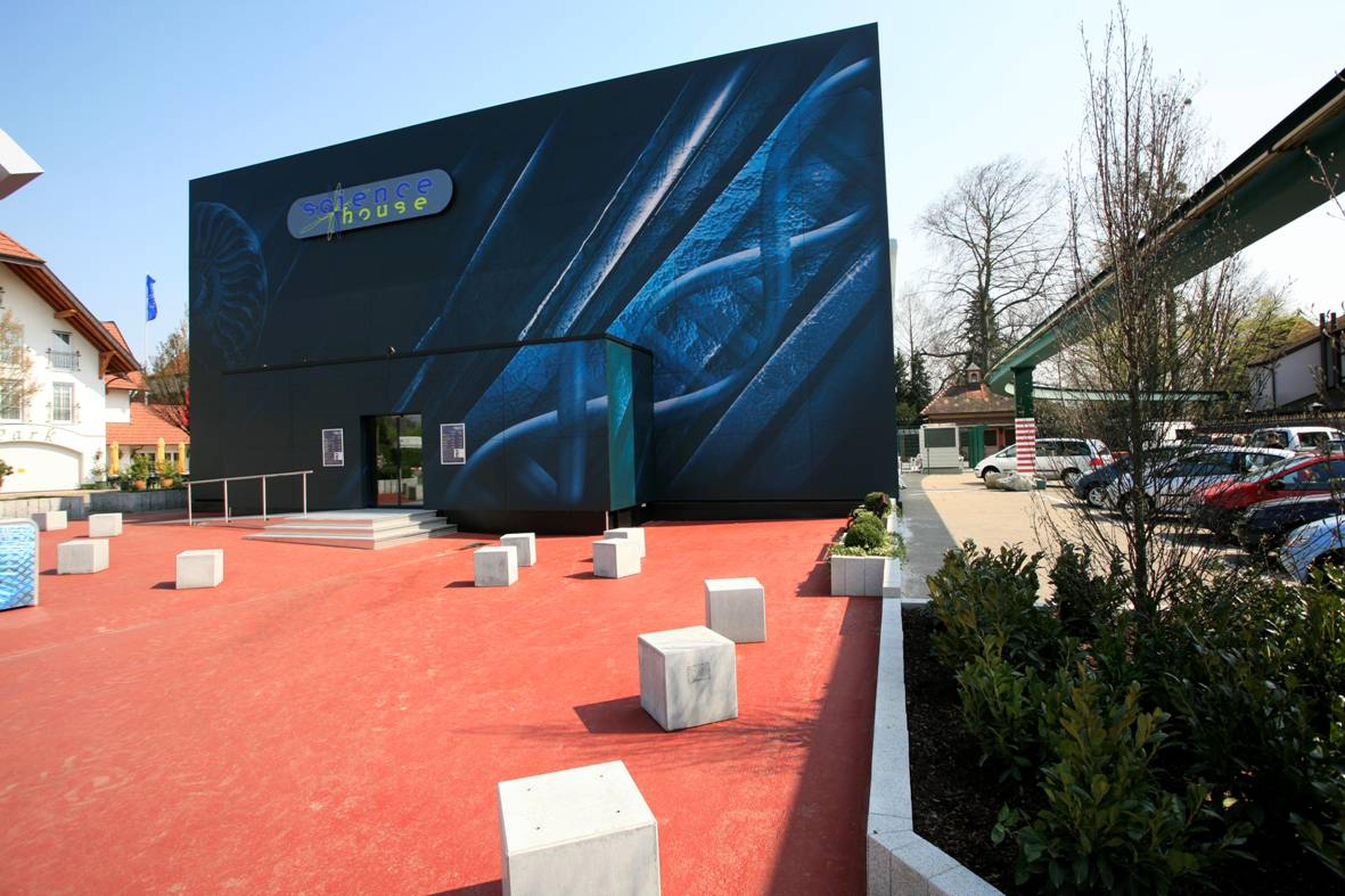 Ein modernes Gebäude mit der Aufschrift „Science House“ und abstraktem Design, das auf einem roten Pflaster aus Betonblöcken steht und von Grünflächen und geparkten Autos umgeben ist.