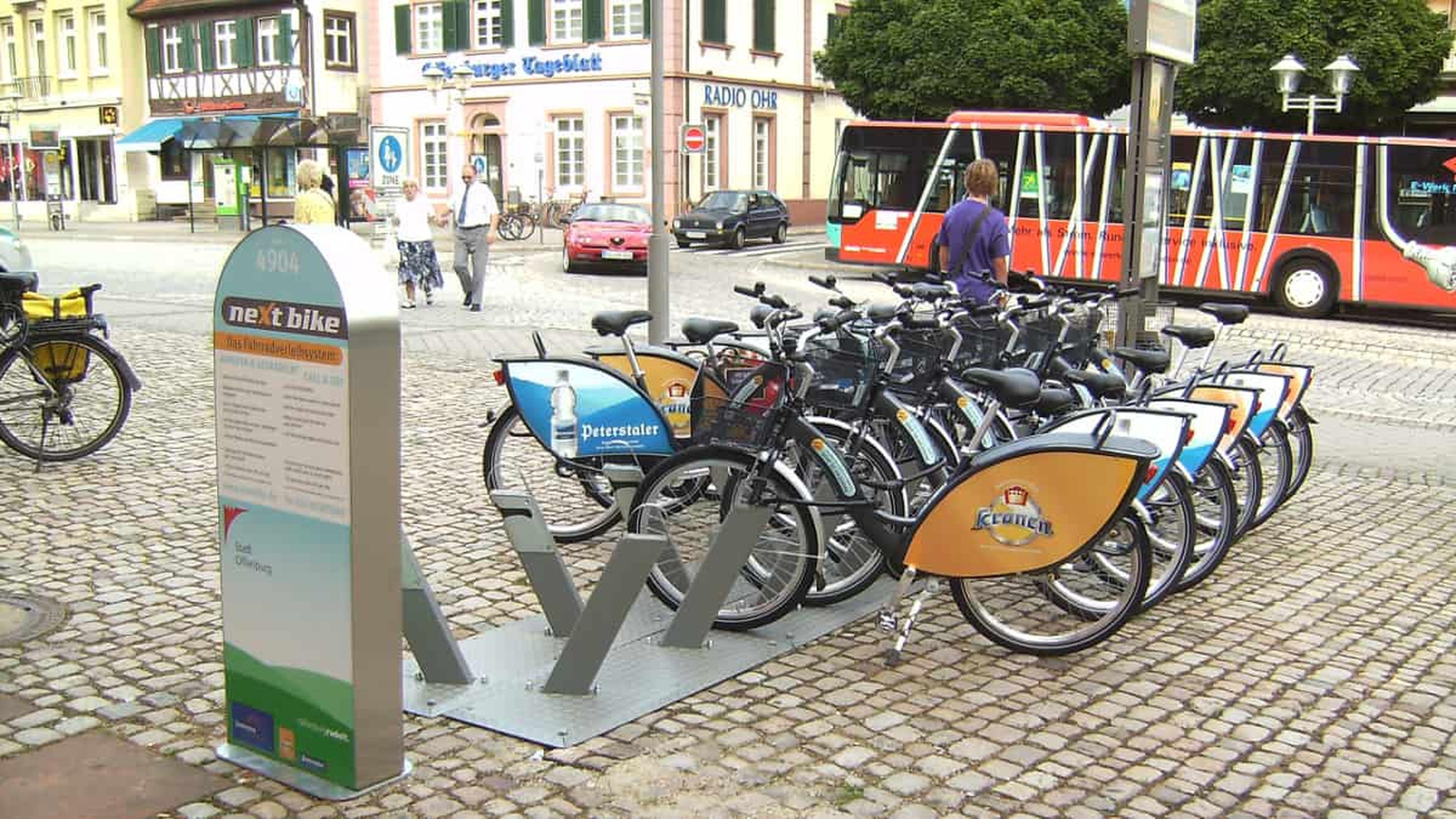 Eine Fahrradverleihstation mit mehreren abgestellten Fahrraedern auf einer Kopfsteinpflasterstraße, einem roten Bus und historischen Gebaeuden im Hintergrund.