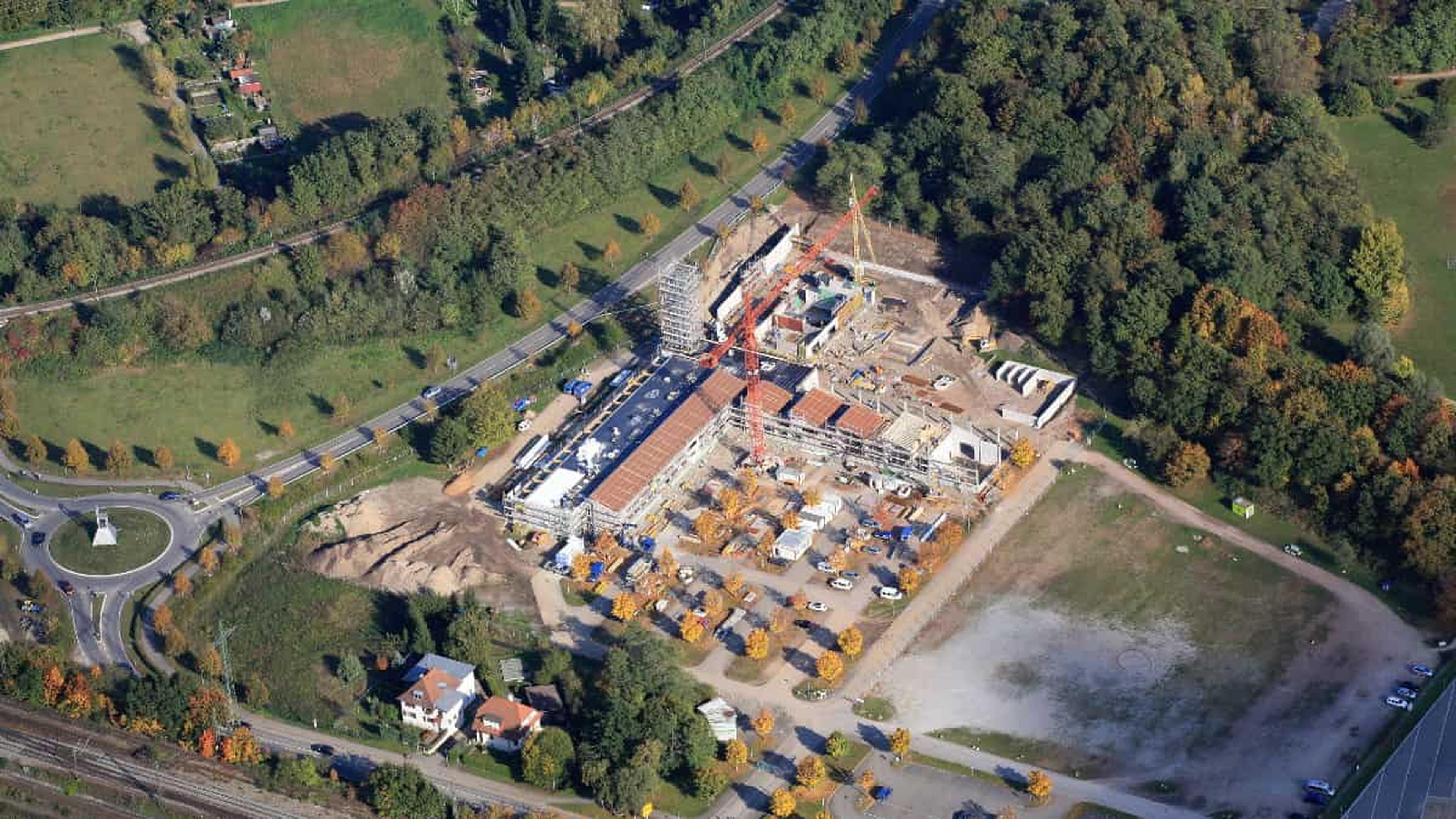 Luftaufnahme einer Baustelle mit Kränen, umgeben von Bäumen und Straßen, in der Nähe eines Kreisverkehrs und vereinzelter Gebäude.