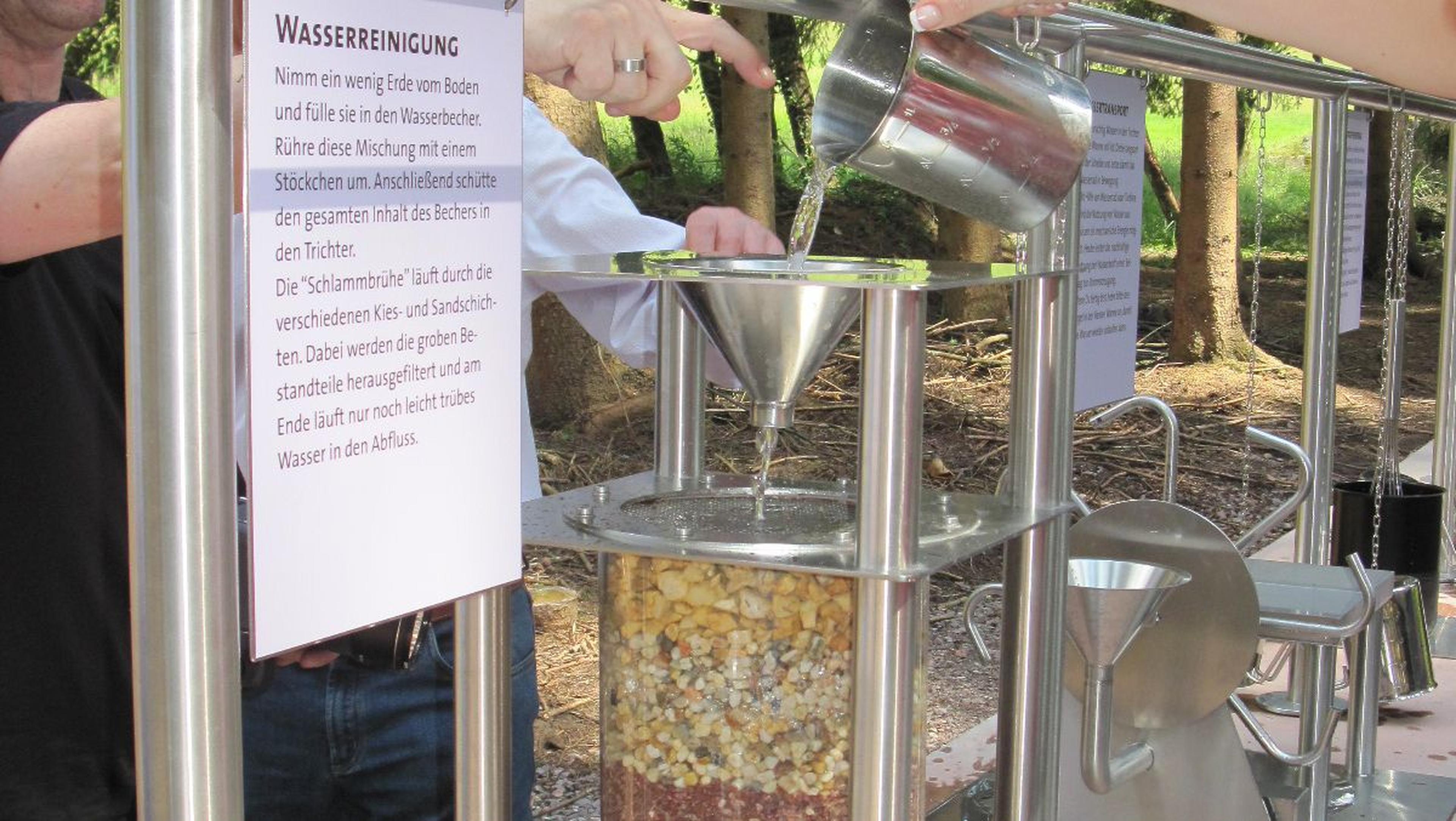 Demonstration der Wasserfilterung im Freien, bei der Wasser mit den Haenden in einen Metalltrichter gegossen wird, der mit Schichten aus Kies und Sand gefuellt ist; Hinweisschild vorhanden.