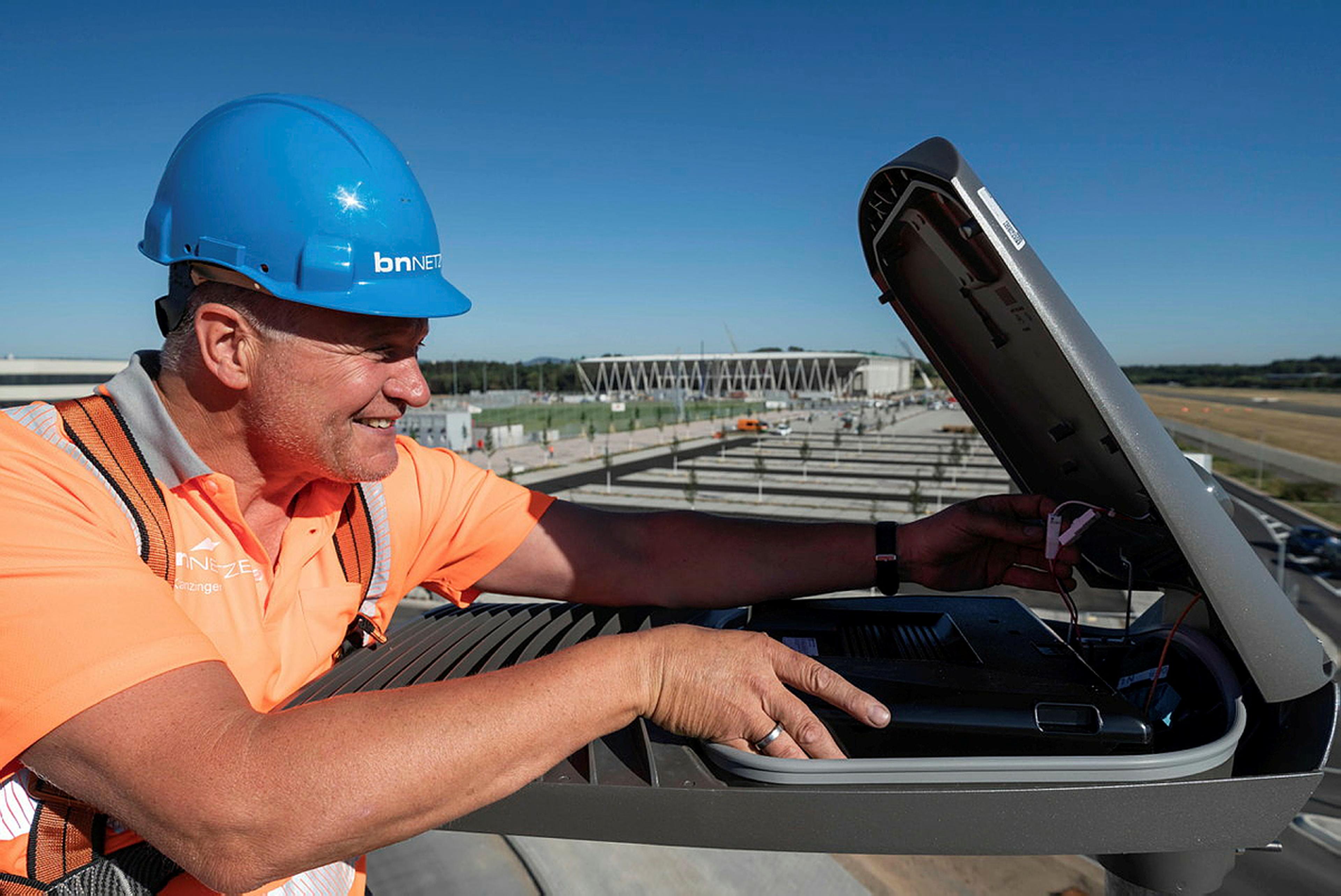 Ein Arbeiter in einer orangefarbenen Weste und einem blauen Helm installiert Geräte auf einem Dach, im Hintergrund ist unter strahlend blauem Himmel ein Stadion zu sehen.