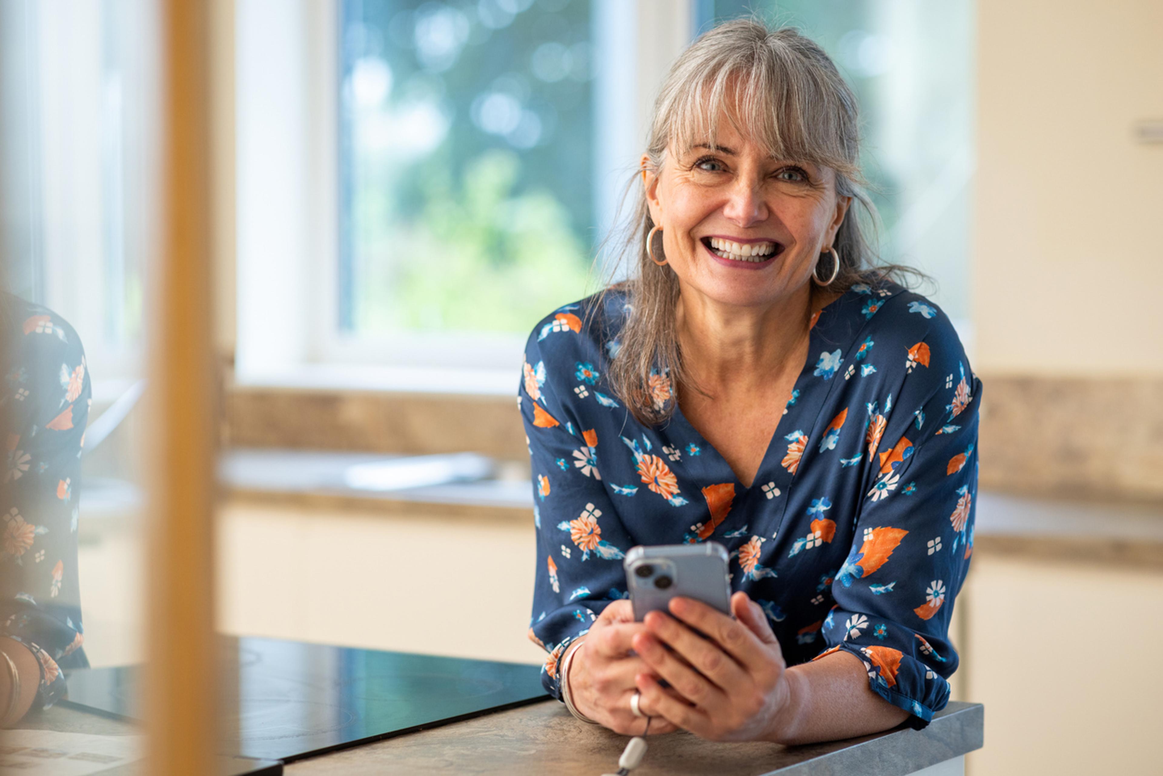 Eine sympathische Frau mit langen grauen Haaren und Pony lächelt selbstbewusst in die Kamera. Sie trägt eine bunte, geblümte Bluse und lehnt an einen Tisch. Sie hält ein Handy in der Hand.