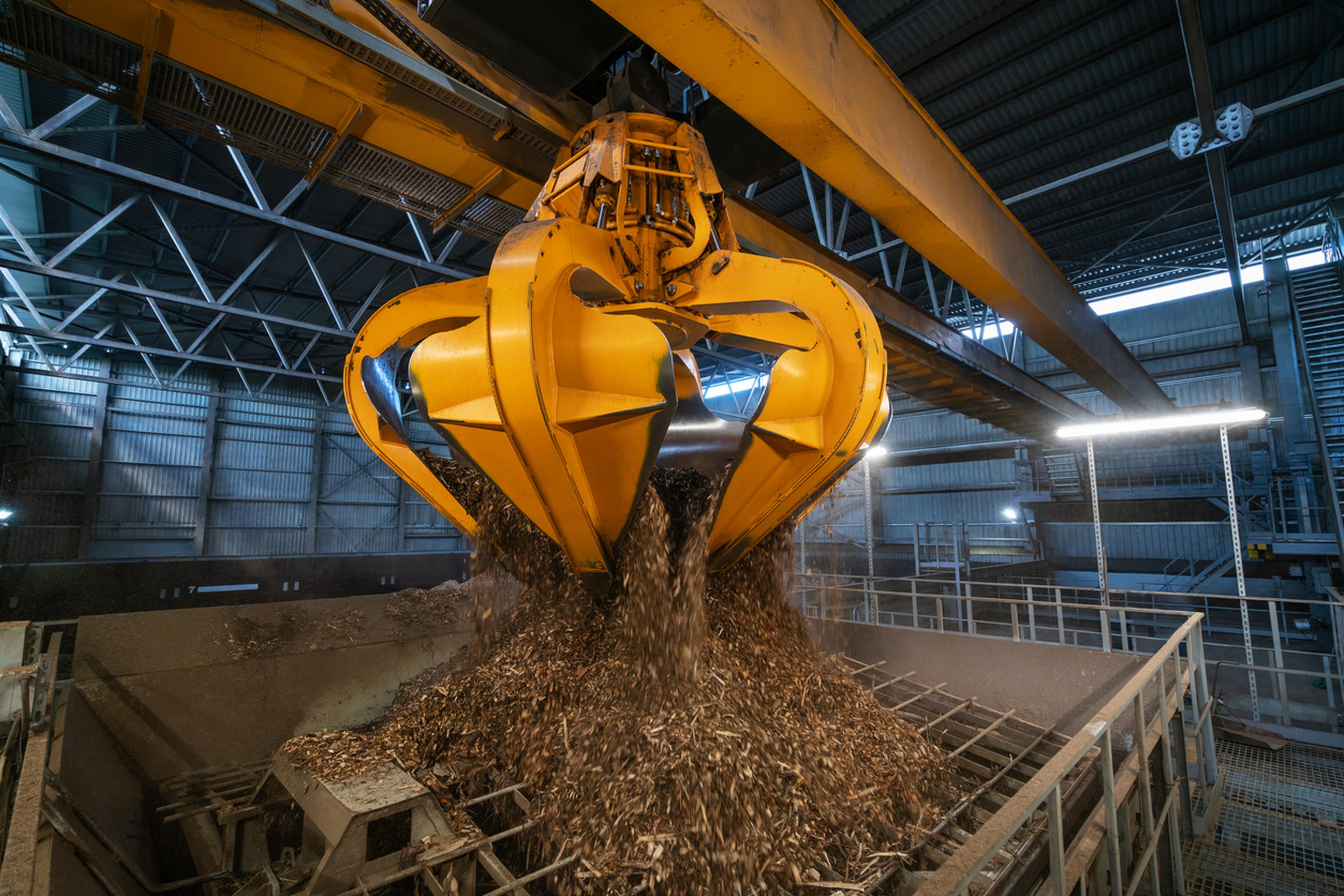 Eine große gelbe Industrie-Greifmaschine lädt Holzspäne auf einen Haufen in einem geräumigen, gut beleuchteten Lagerhaus..