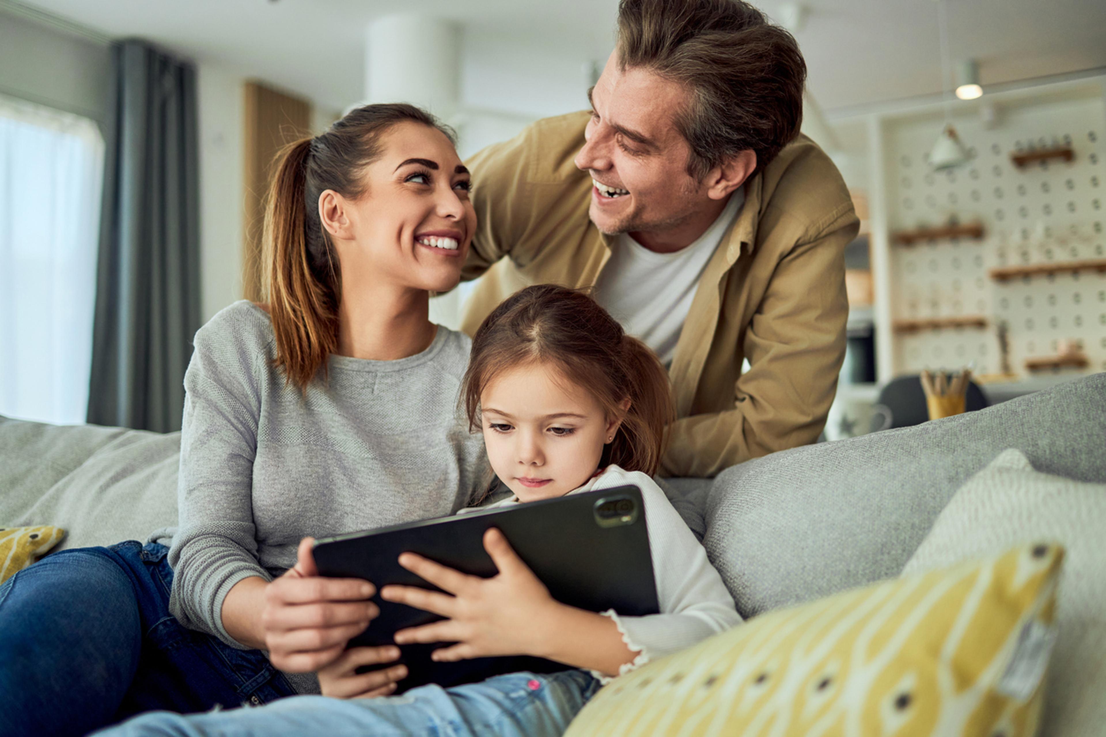 Die Eltern lächeln, während ihre kleine Tochter auf dem Sofa konzentriert auf ein Tablet schaut.